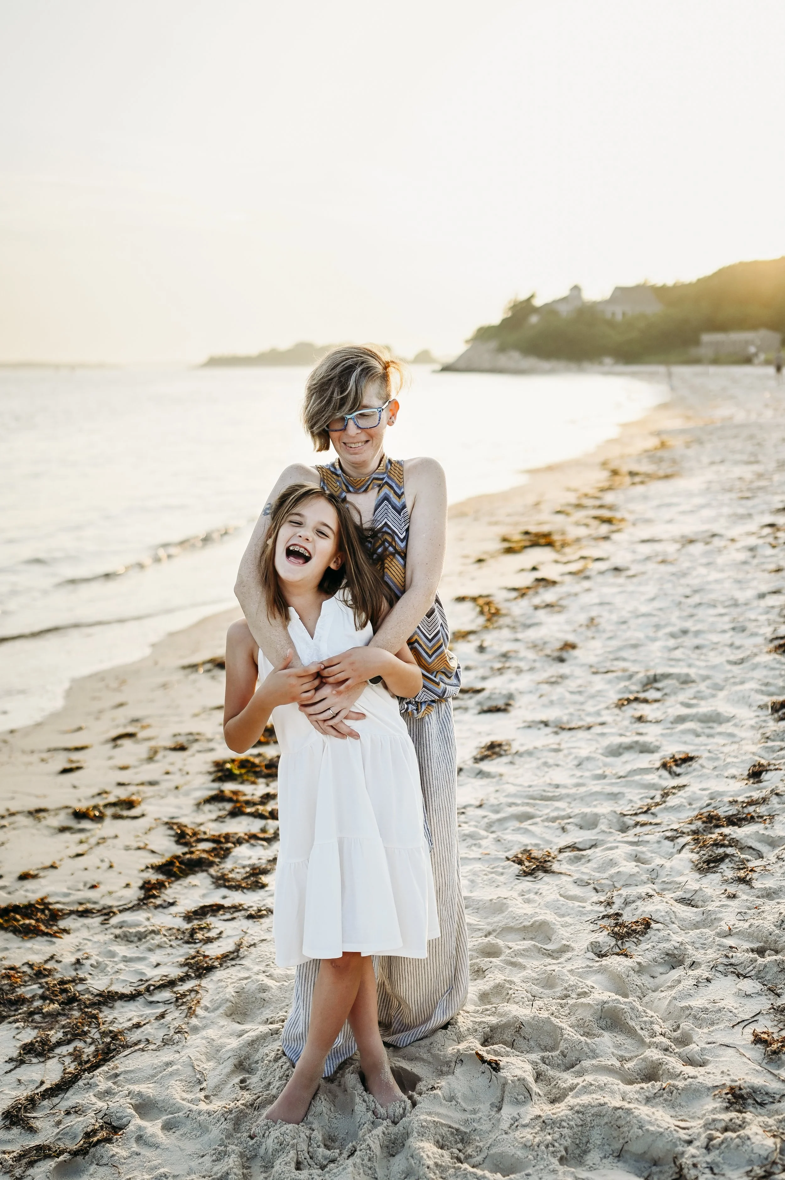A woman and a young girl laughing and hugging on the beach during sunset.