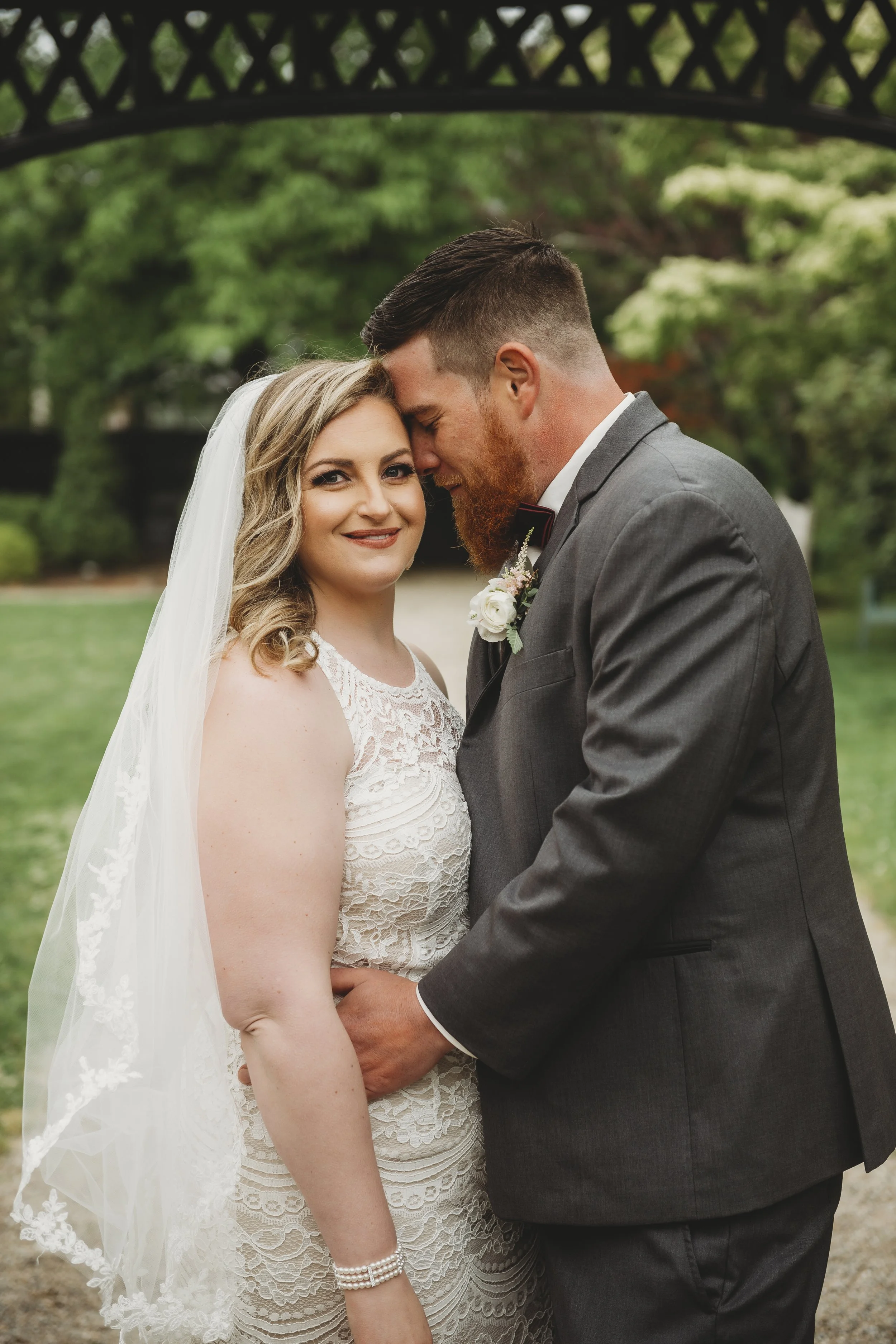 A bride and groom on their wedding day, standing close together under a decorative arch, outdoors with green trees in the background. The bride has blonde hair, wears a lace wedding dress and a veil, and is smiling at the camera. The groom has a bear