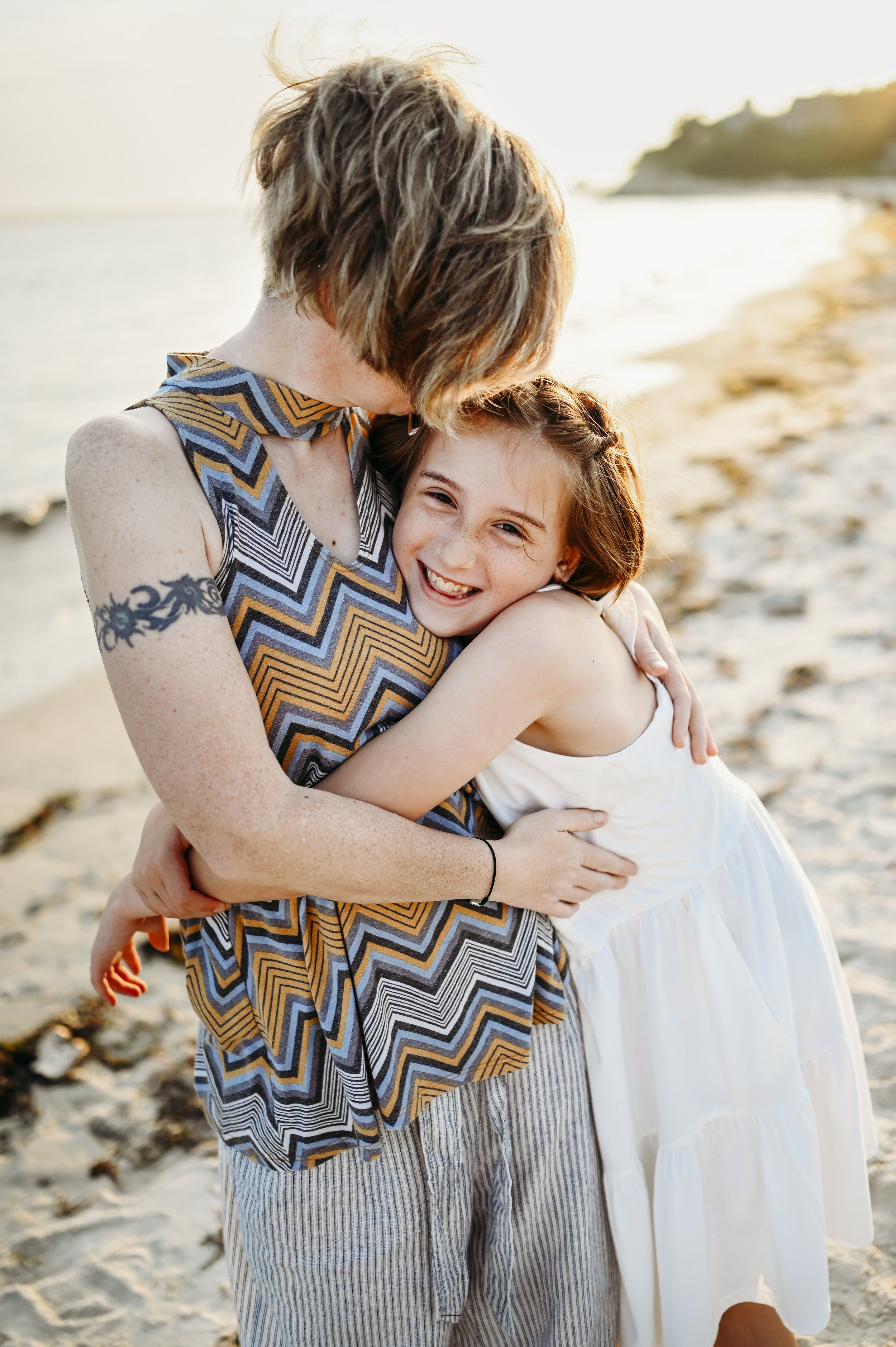 A woman and a young girl hugging each other on the beach during sunset, smiling happily.