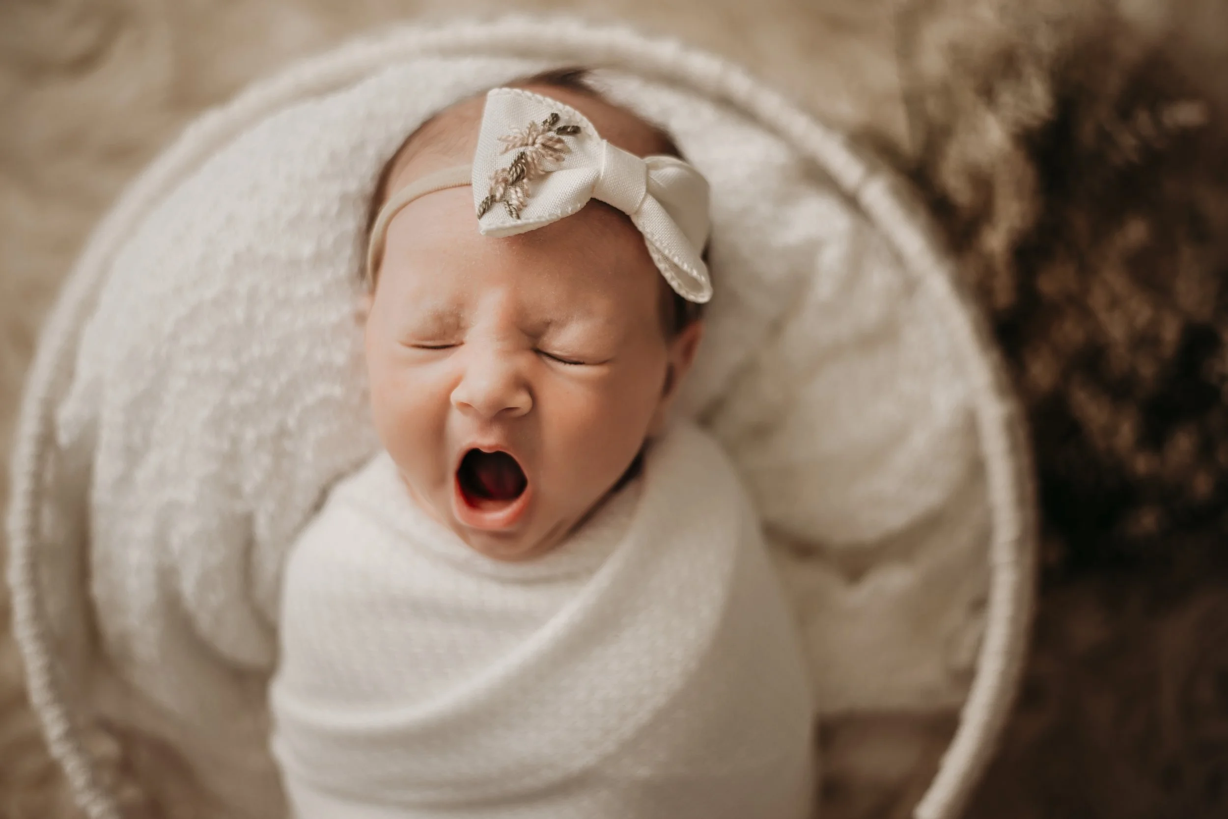 A newborn baby yawning while swaddled in a white blanket, wearing a decorative headband with a bow.