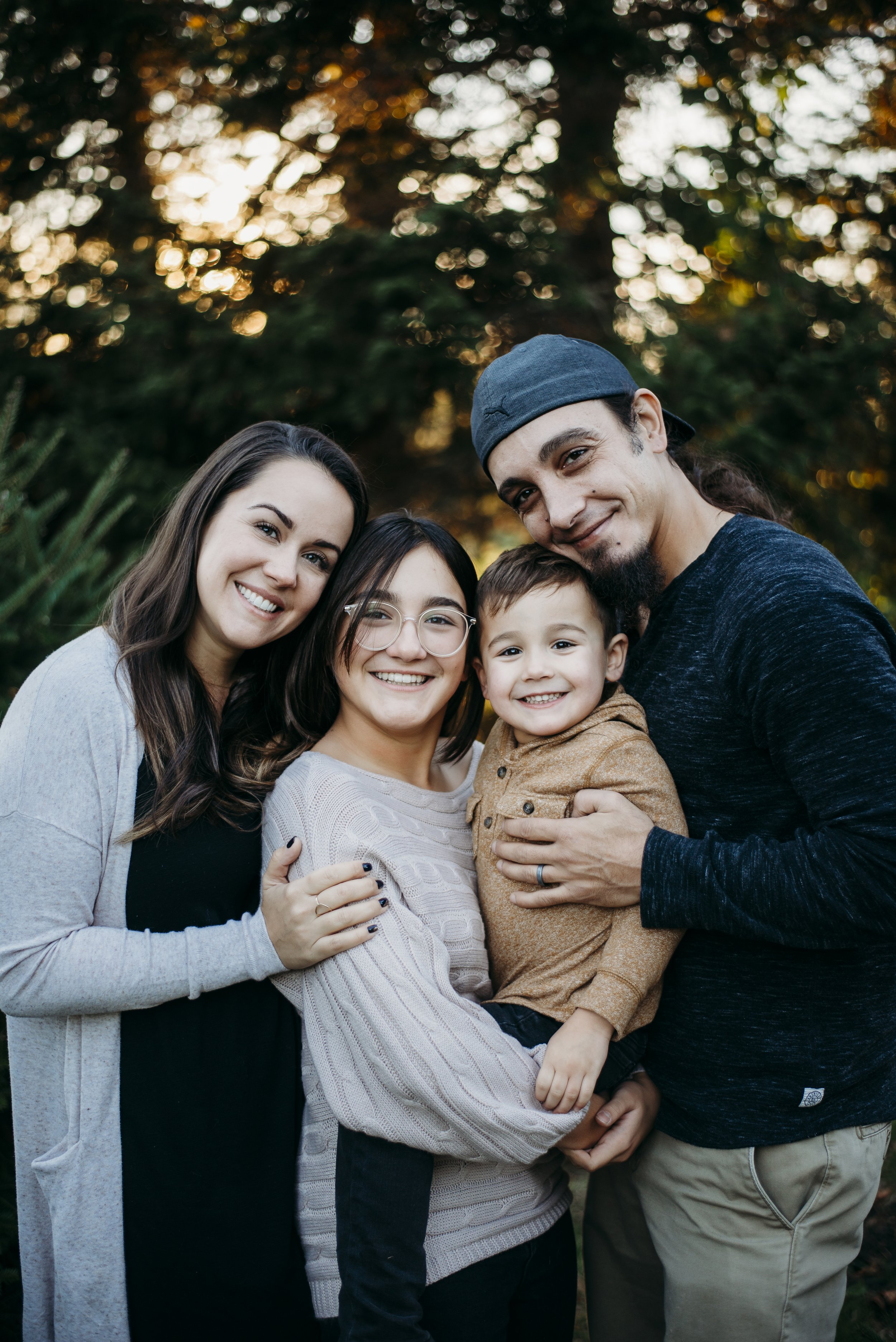 A smiling diverse family of four, including two women, one man, and a young boy, sharing a hug outdoors during sunset.