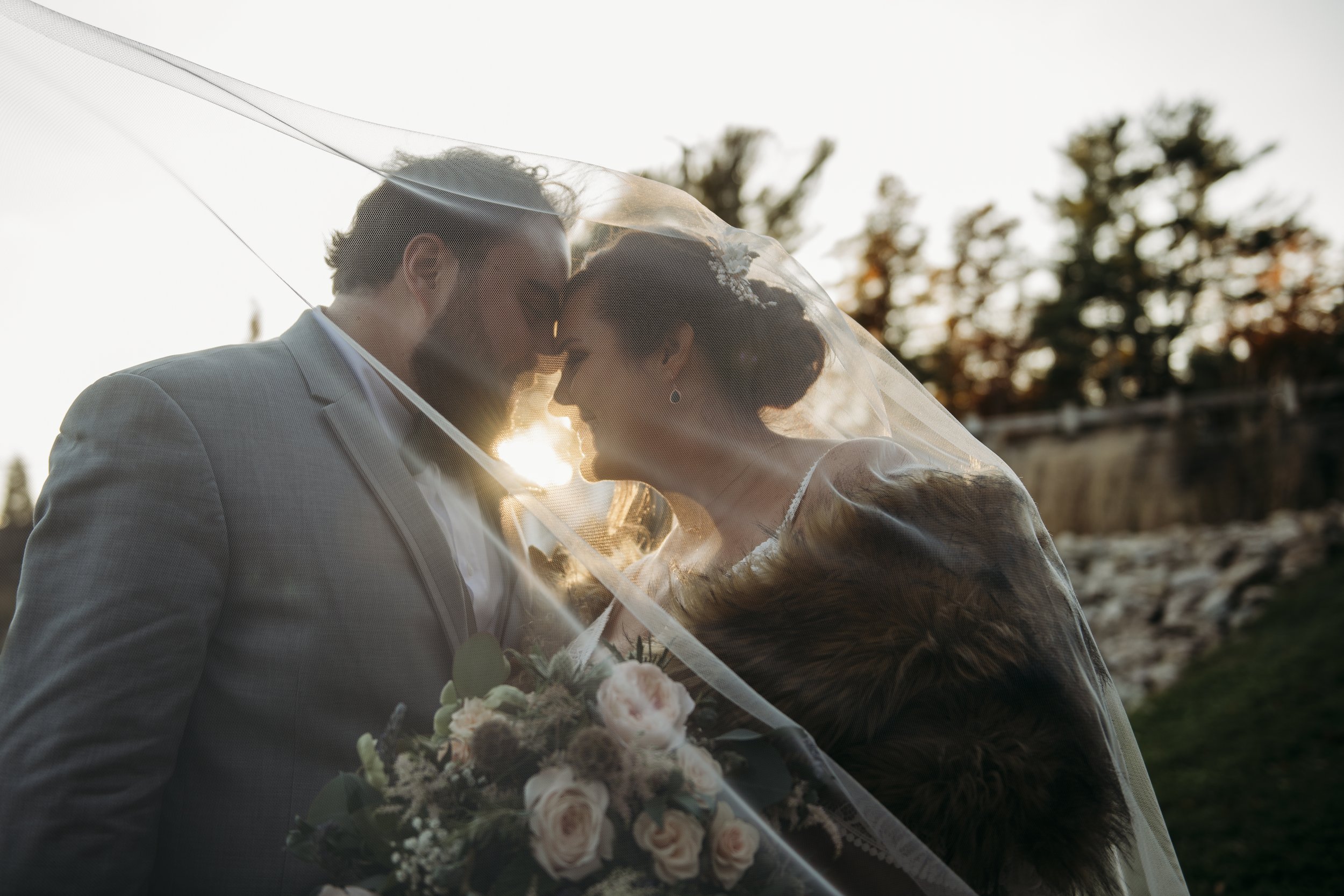A bride and groom nose-to-nose under a veil, backlit by the setting sun, outdoors with trees in the background.