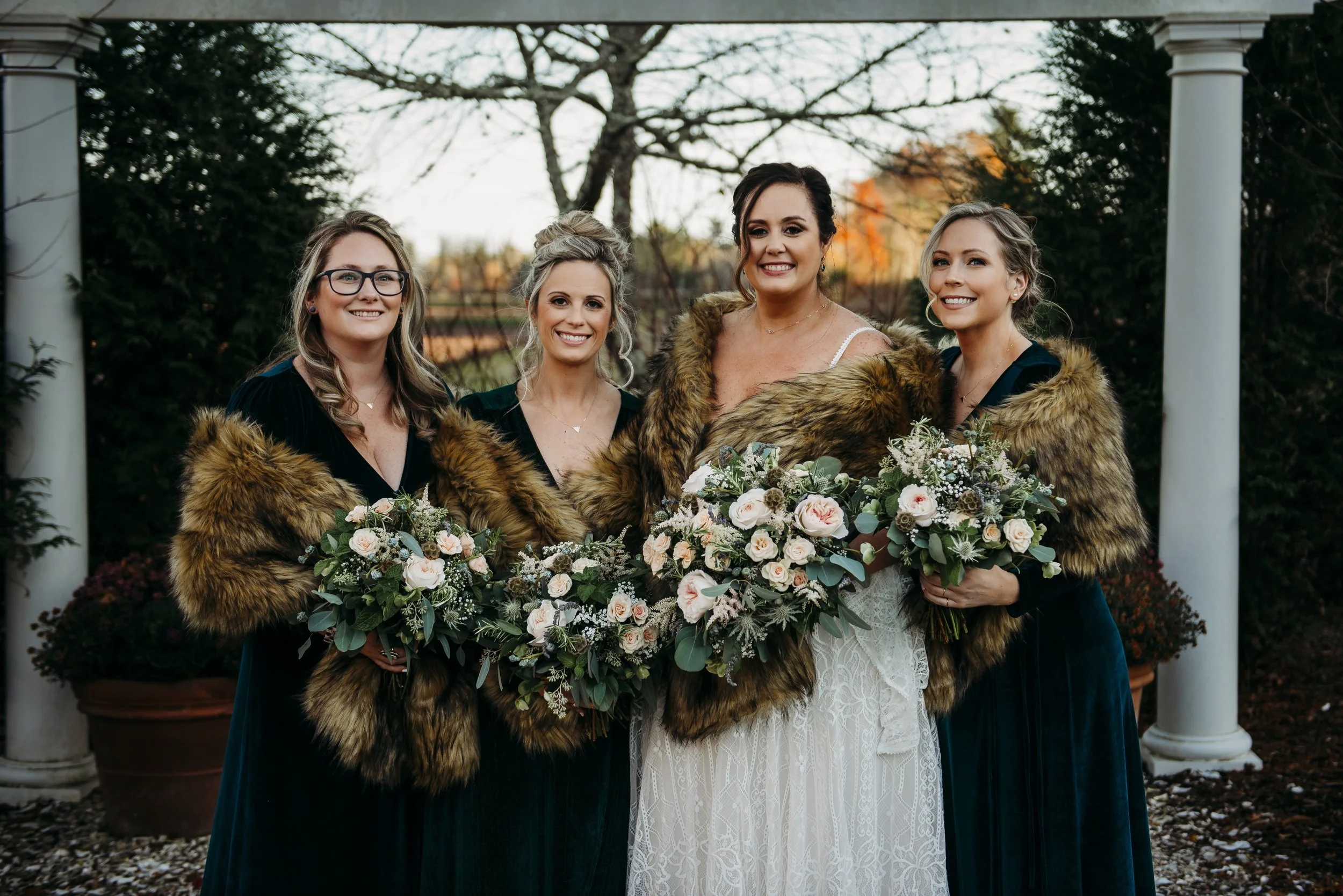 A bride and her bridesmaids standing outdoors, all wearing fur shawls, holding bouquets of flowers, with trees and columns in the background.