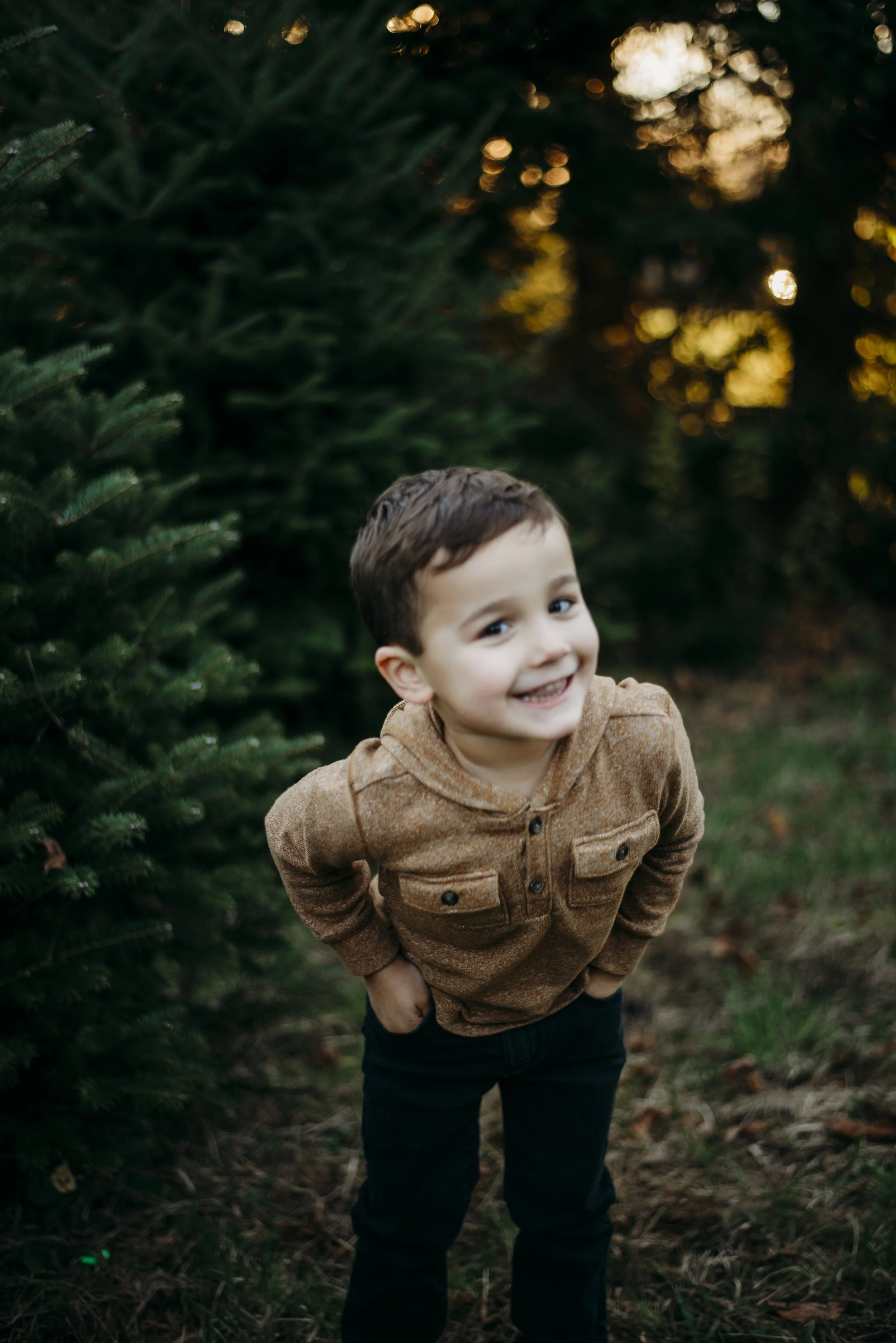 A smiling young boy with short dark hair, wearing a brown hoodie and black pants, standing outdoors between Christmas trees during sunset.