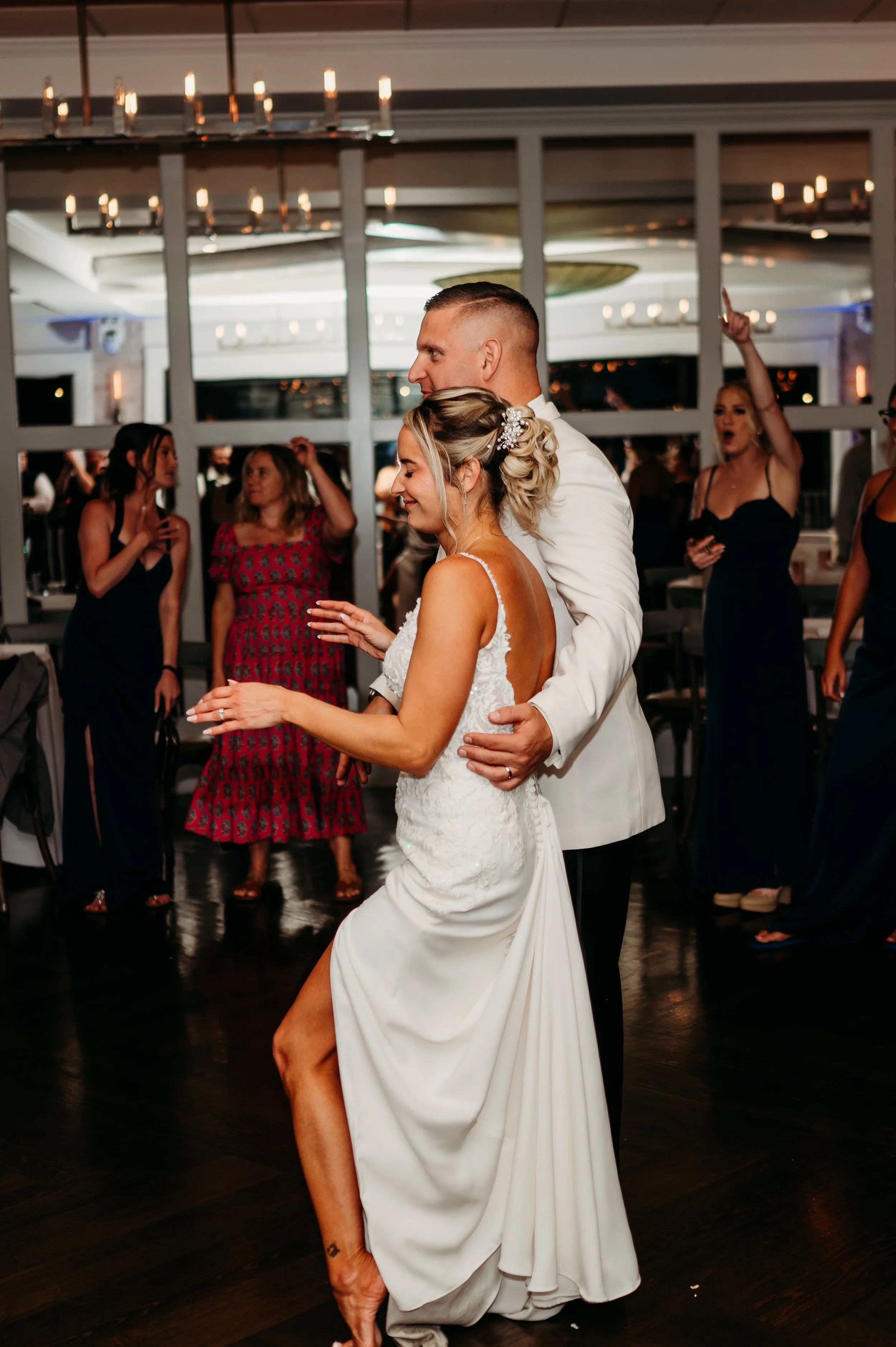Couple dancing at a wedding reception, surrounded by guests in formal attire, in a dimly lit indoor venue with chandeliers.