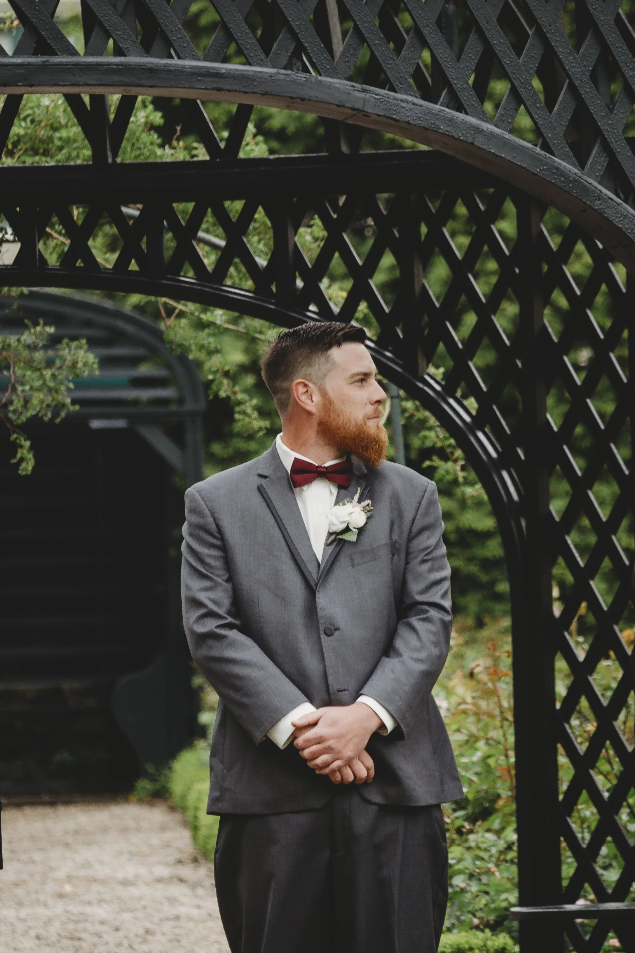 A man dressed in a gray suit with a white shirt, burgundy bow tie, and boutonniere, standing outdoors near a black wooden lattice arch, with green foliage in the background.