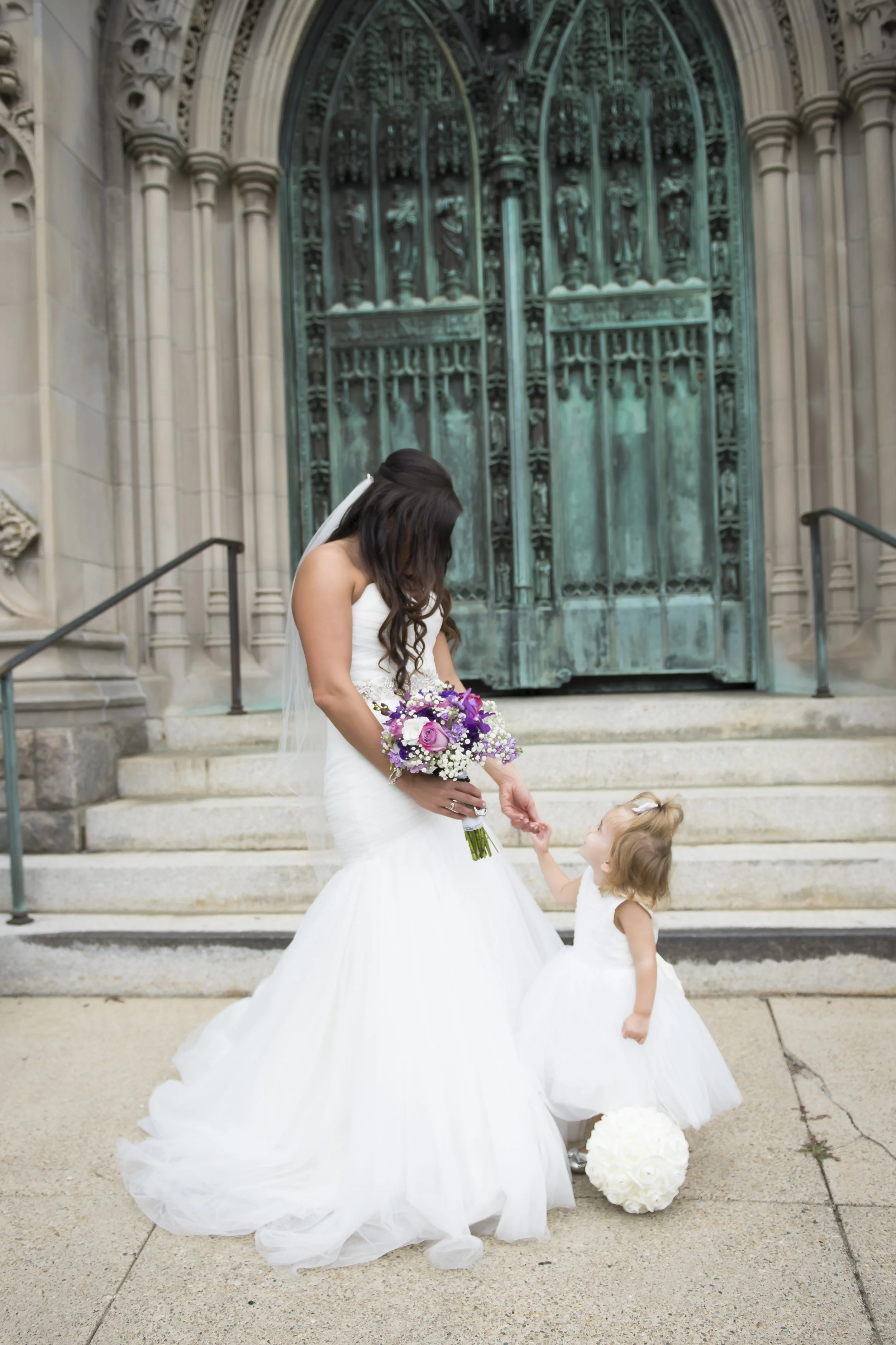 A bride in a white wedding gown holding a bouquet of purple and white flowers, gently touching the hand of a young girl in a white dress, standing on steps outside a building with ornate green doors.