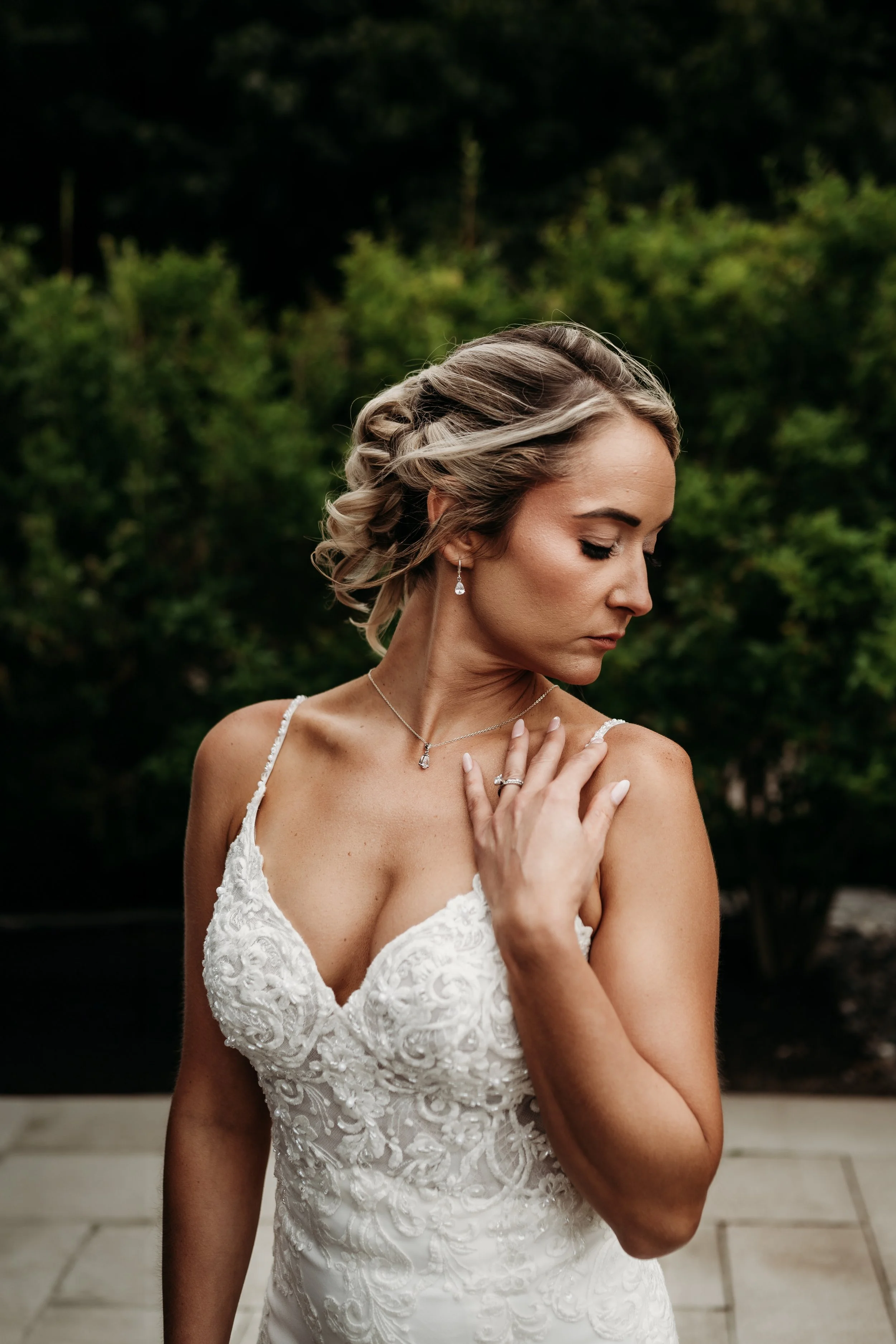 Bride with blonde, curly hair in a white lace wedding dress, wearing jewelry, standing outdoors with greenery in the background.
