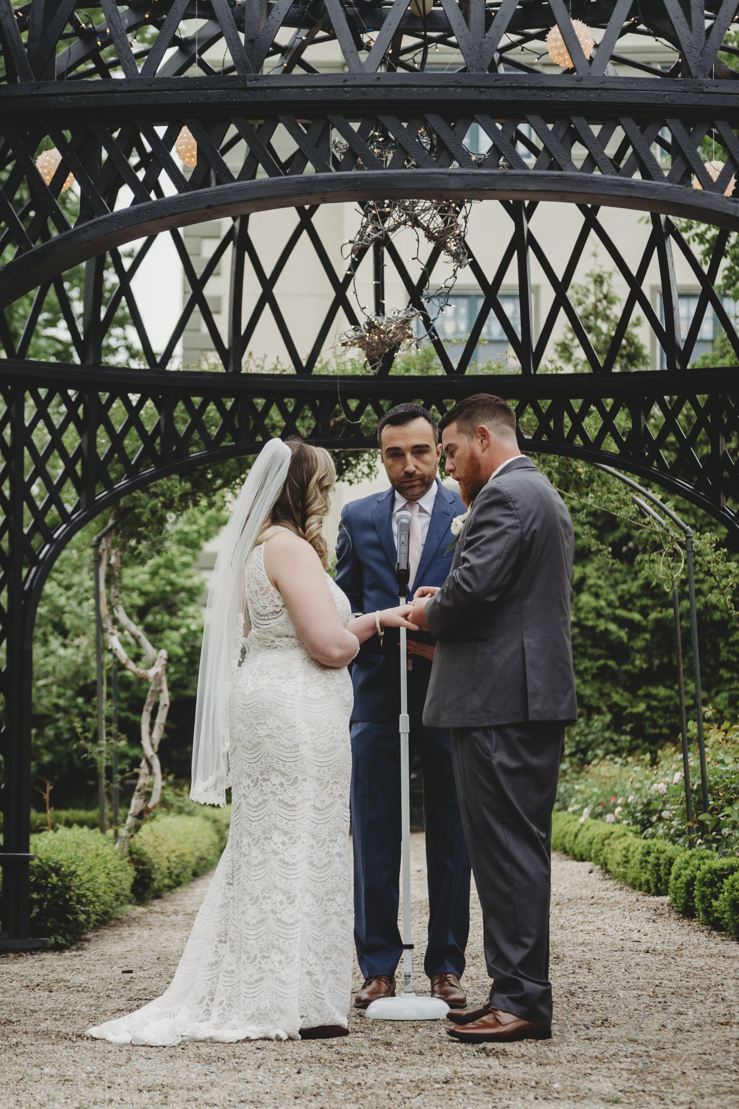 Couple getting married outdoors under a black lattice arch with greenery in the background, officiant standing between them.