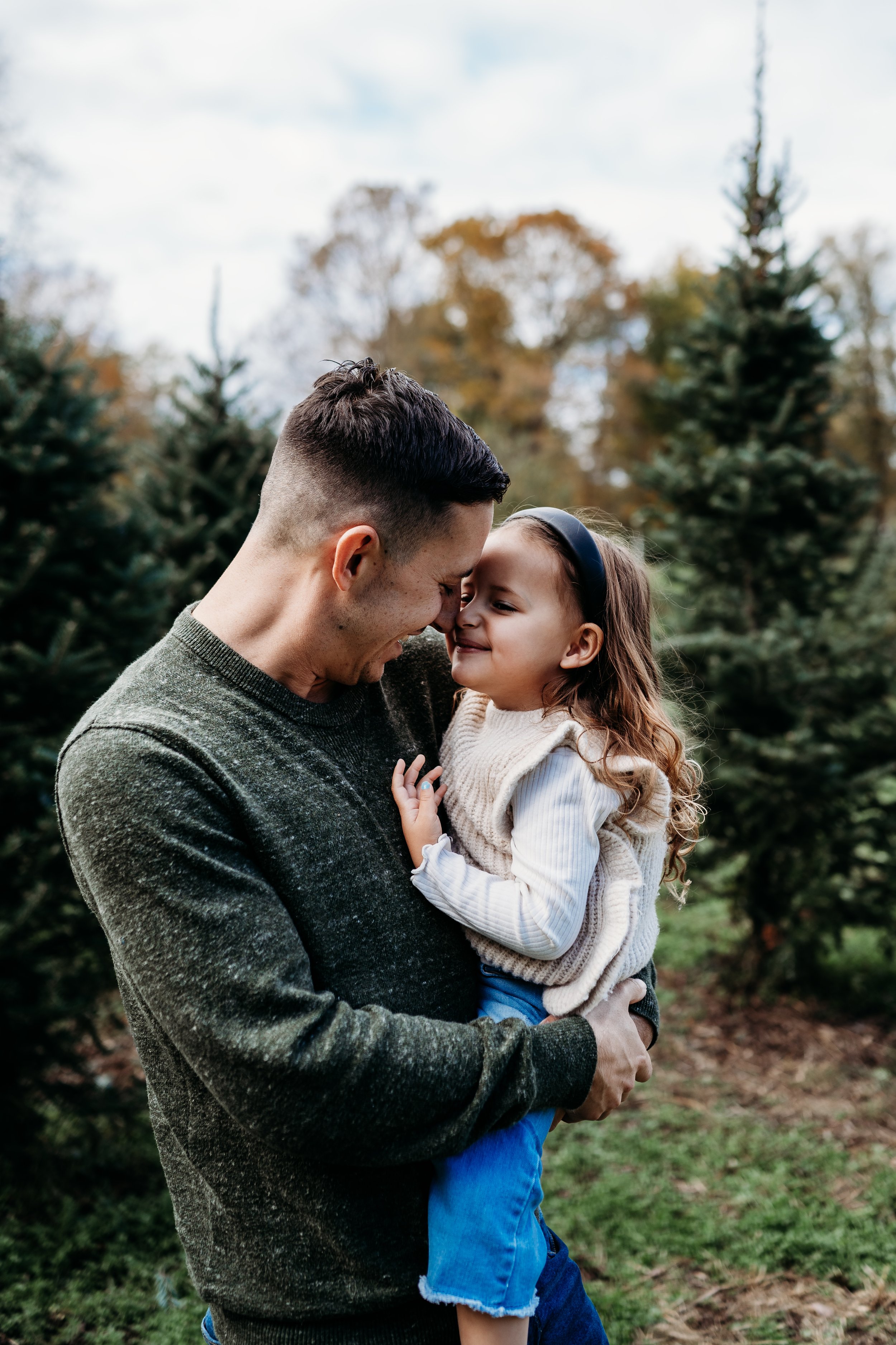 A man holding a young girl outdoors among Christmas trees, touching foreheads, smiling, in a park with fall-colored trees in the background.