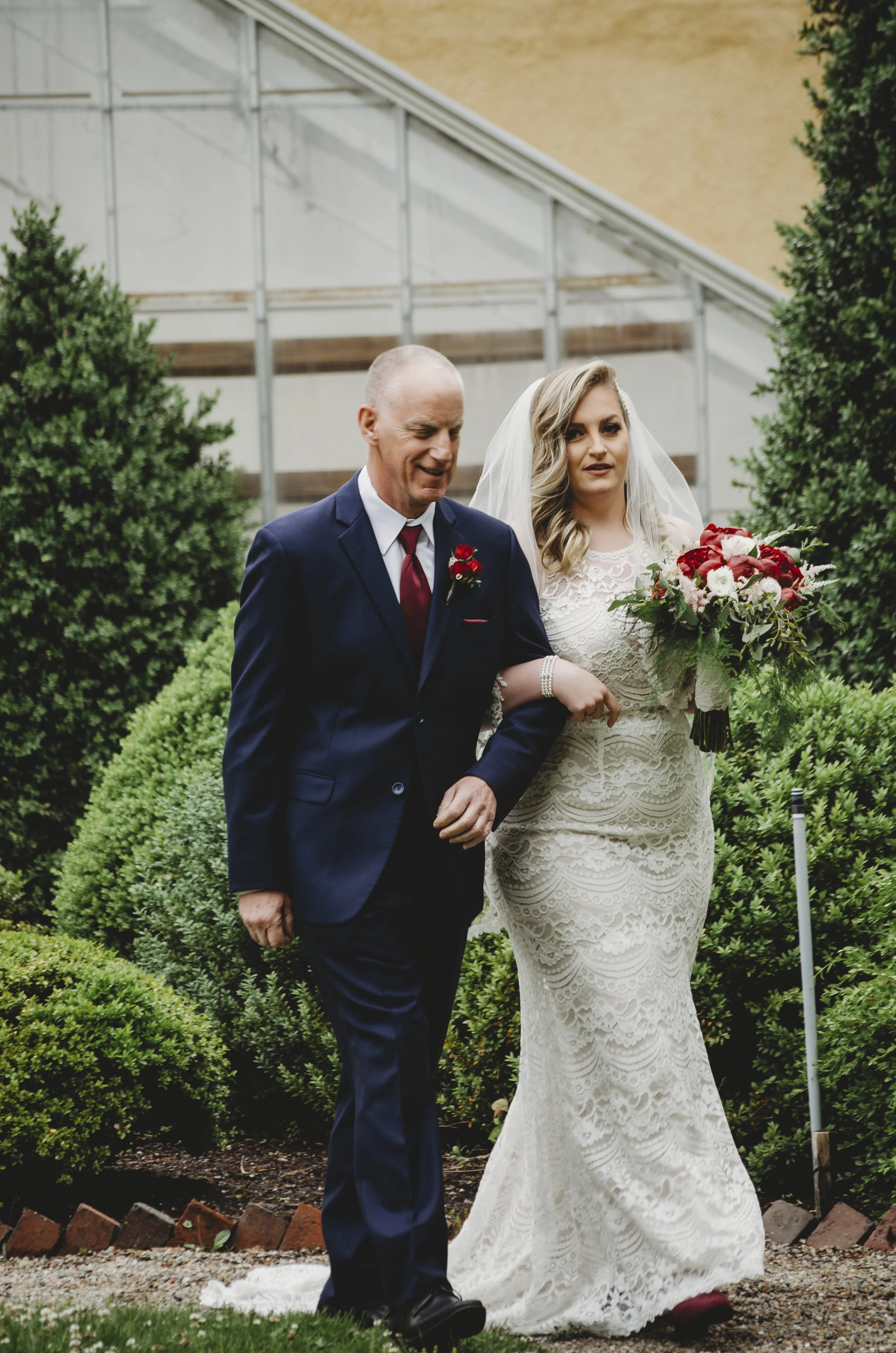 A bride with blonde wavy hair holding a bouquet of red and white flowers, walking arm-in-arm with an older man in a dark suit during a wedding ceremony outdoors.