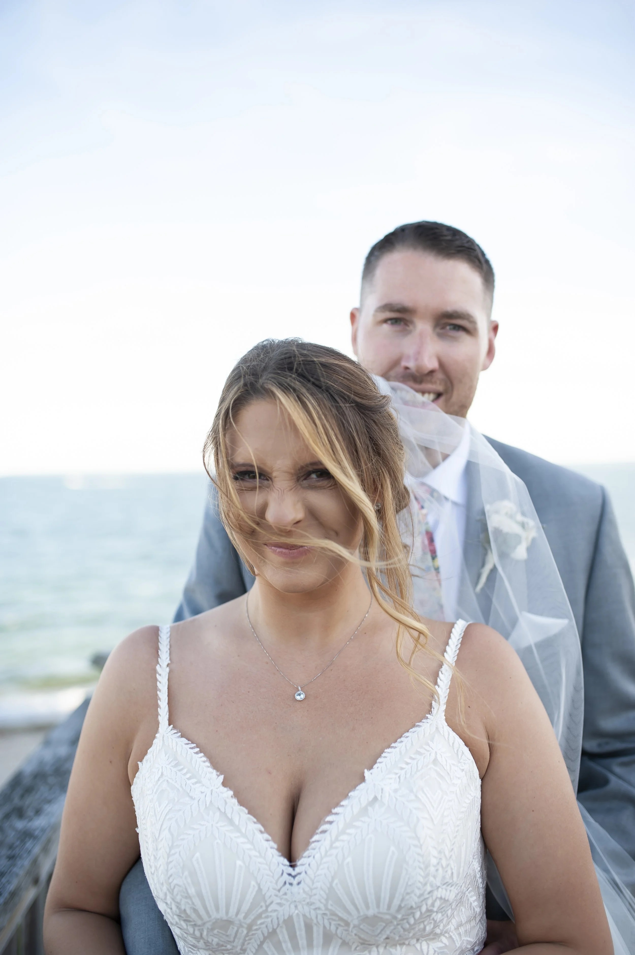 A bride and groom on a beach, with the bride making a playful face and the groom smiling, standing in front of the ocean.