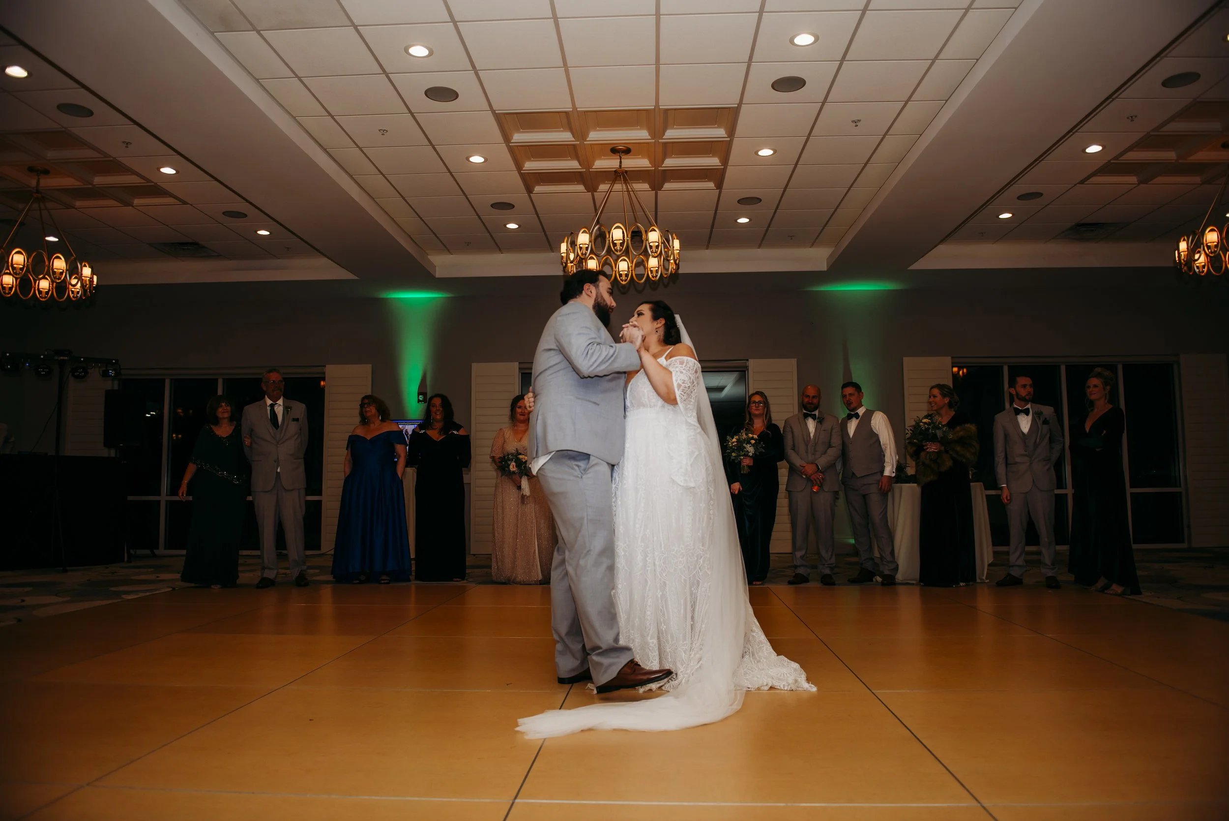 A newlywed couple dancing during their wedding reception with guests watching in a ballroom under warm lighting and chandeliers.