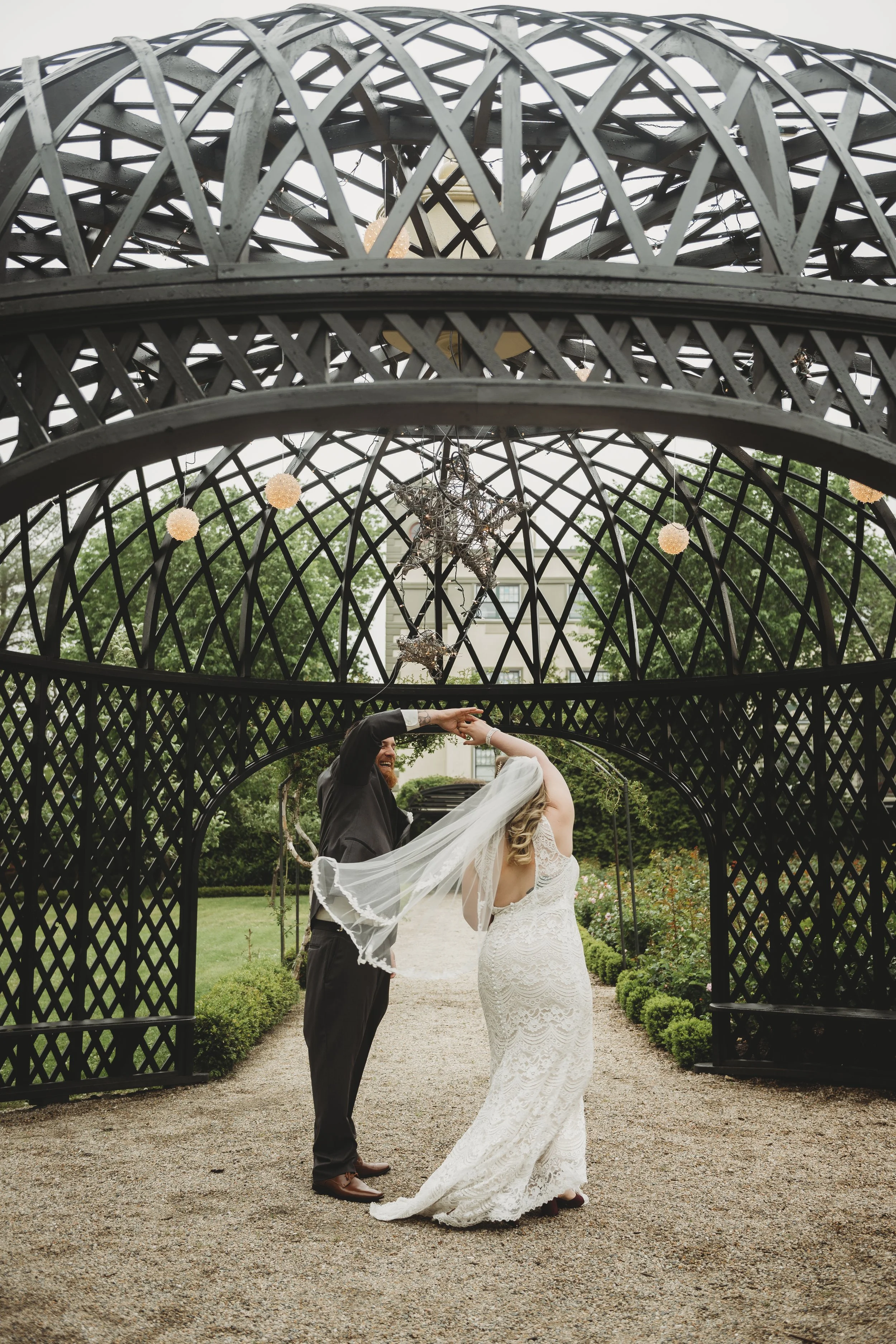 A bride and groom dancing under a decorative black lattice gazebo with star and spherical light decorations, surrounded by greenery.