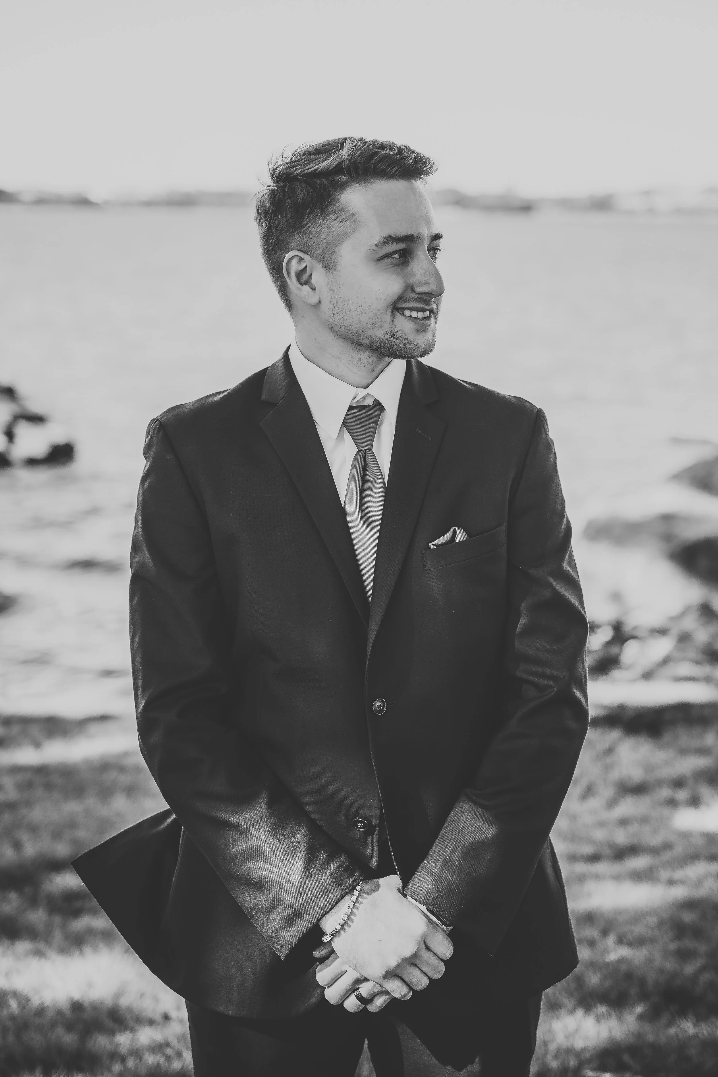 Black and white photo of a young man in a suit, standing outdoors near water, smiling and looking to the right.