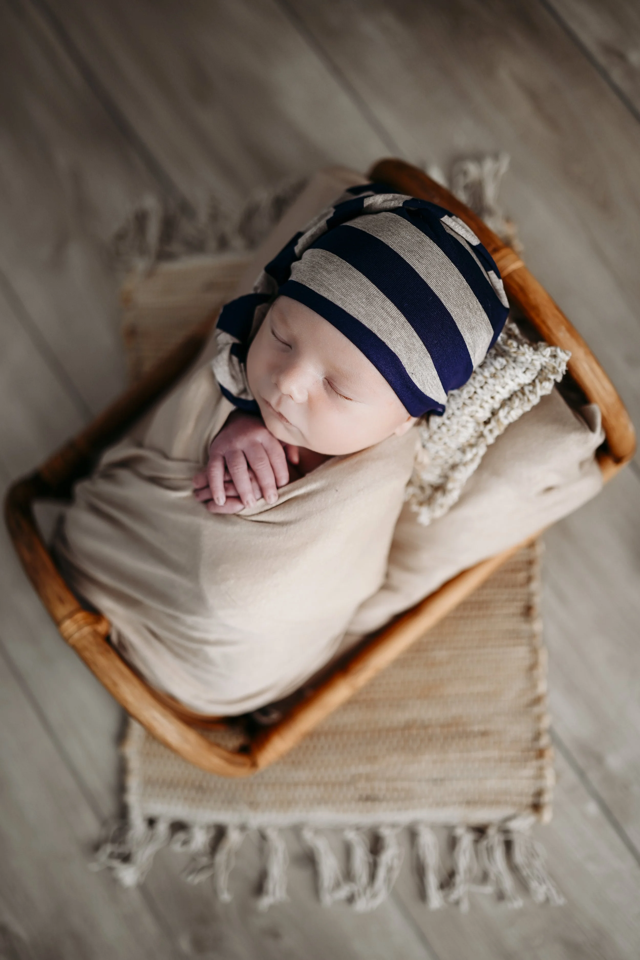 A sleeping baby wrapped in a beige blanket, wearing a navy blue and gray striped hat, lying on a small bed inside a wicker basket, on a woven rug.