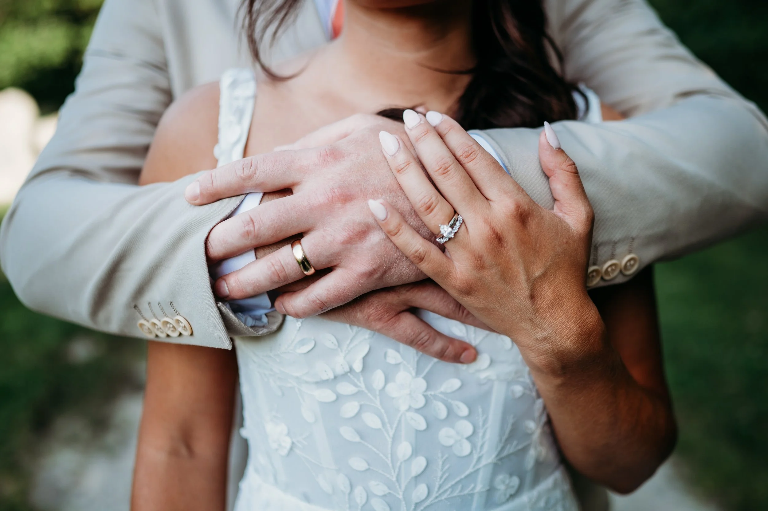 Close-up of a man and woman embracing; the man's arms are around the woman's shoulders, and the woman's hands, showing a wedding ring, are resting on the man's arm. The woman is wearing a white wedding dress with embroidery and a diamond engagement r