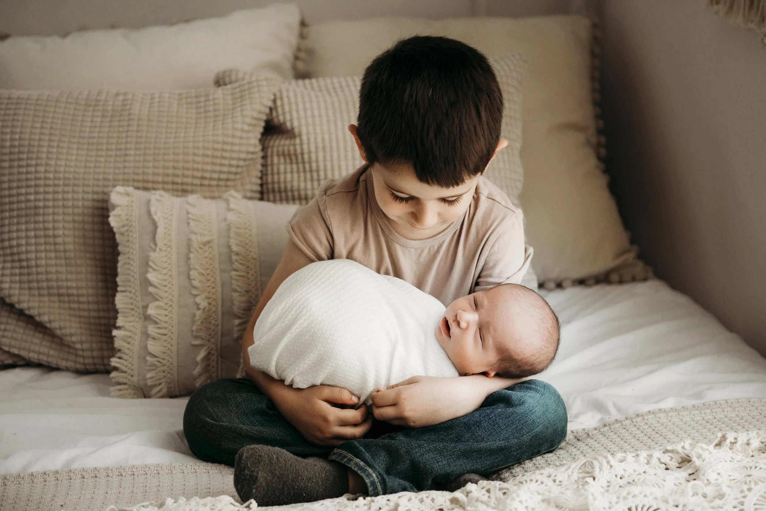 A young boy sitting on a bed holding a newborn baby wrapped in a white blanket, with beige and checkered pillows in the background.