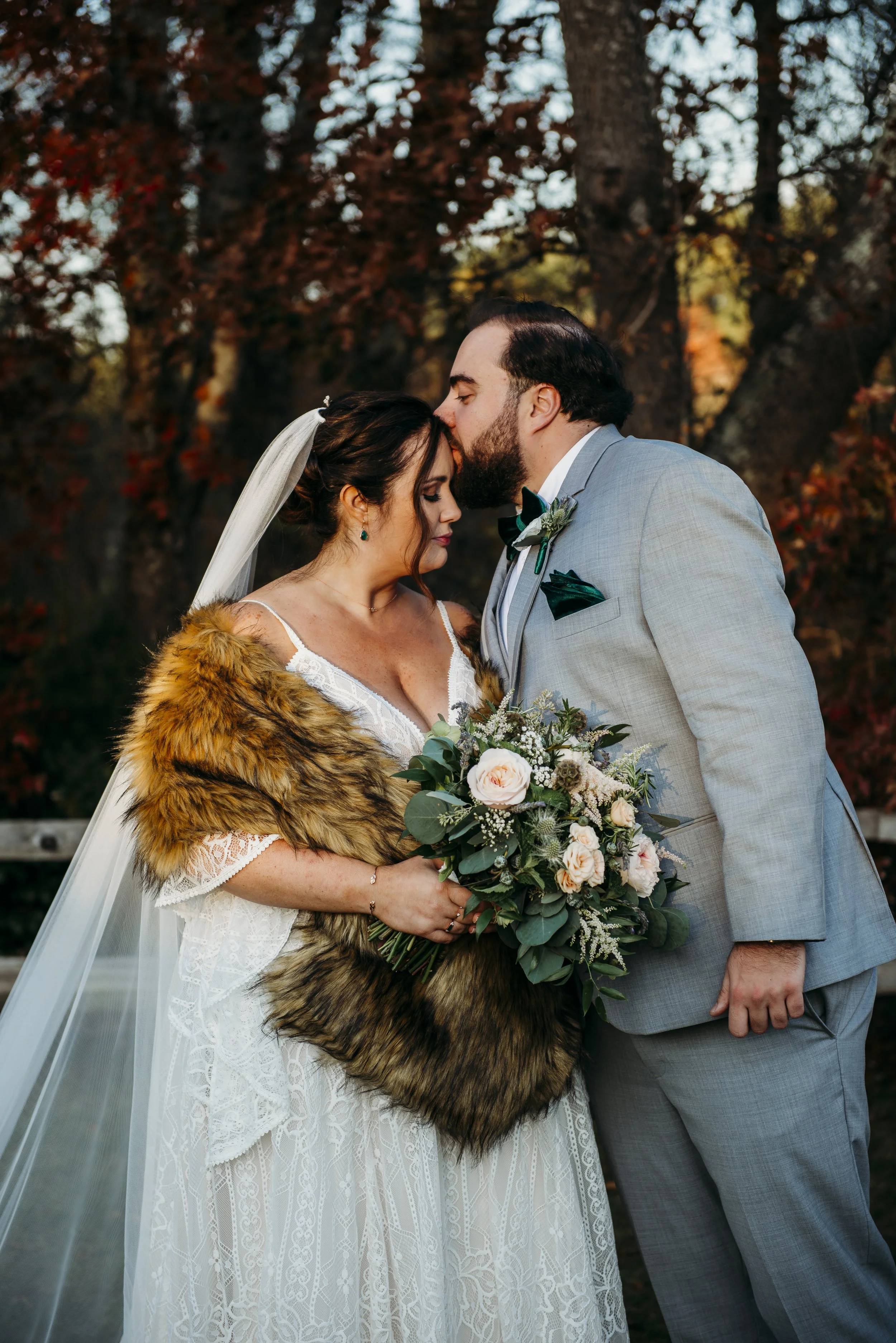A bride and groom sharing an intimate moment outdoors during their wedding, with the groom kissing the bride's forehead, in front of autumn-colored trees.