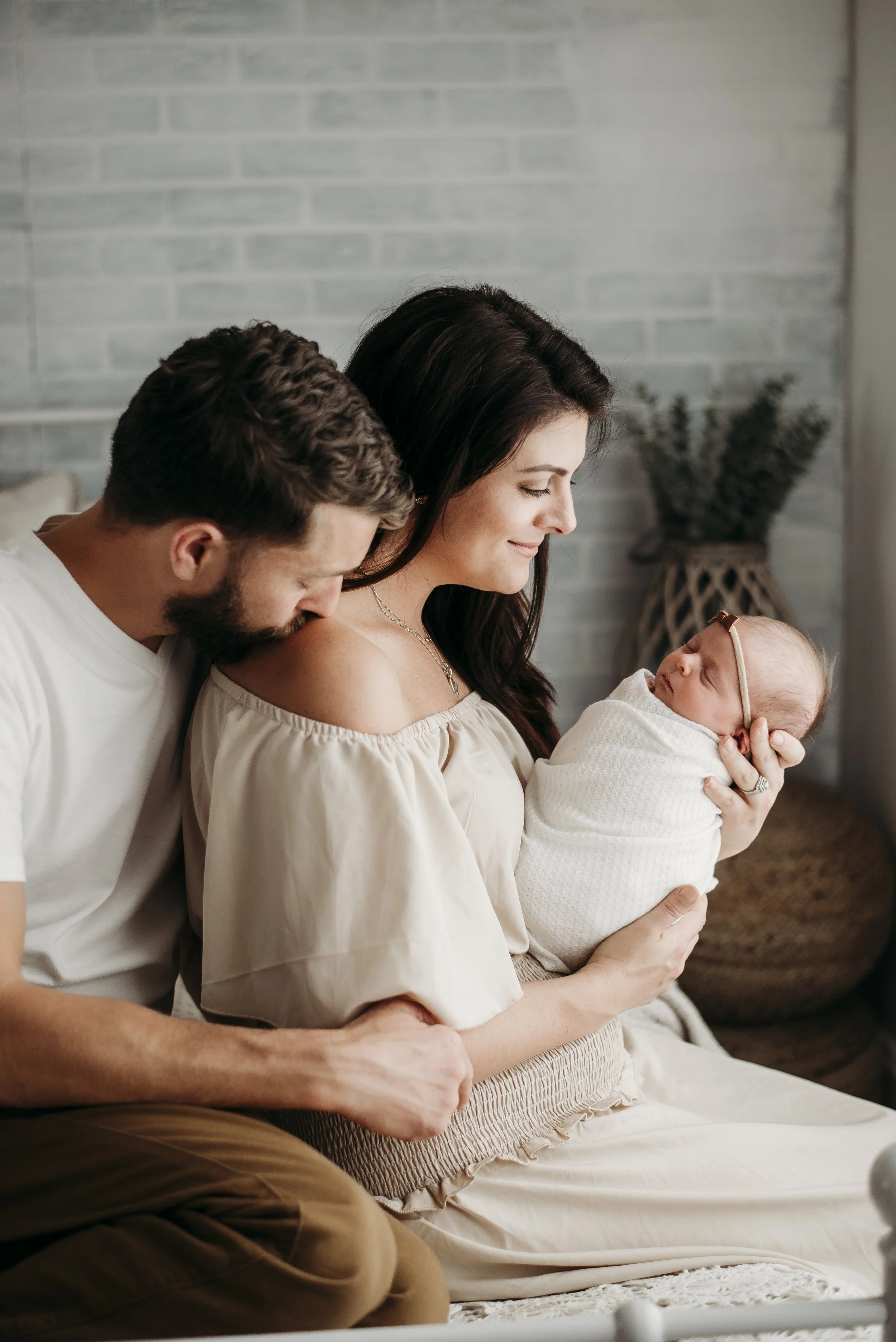A woman holding a newborn baby, with a man leaning in close, they are in a cozy room with a brick wall background.