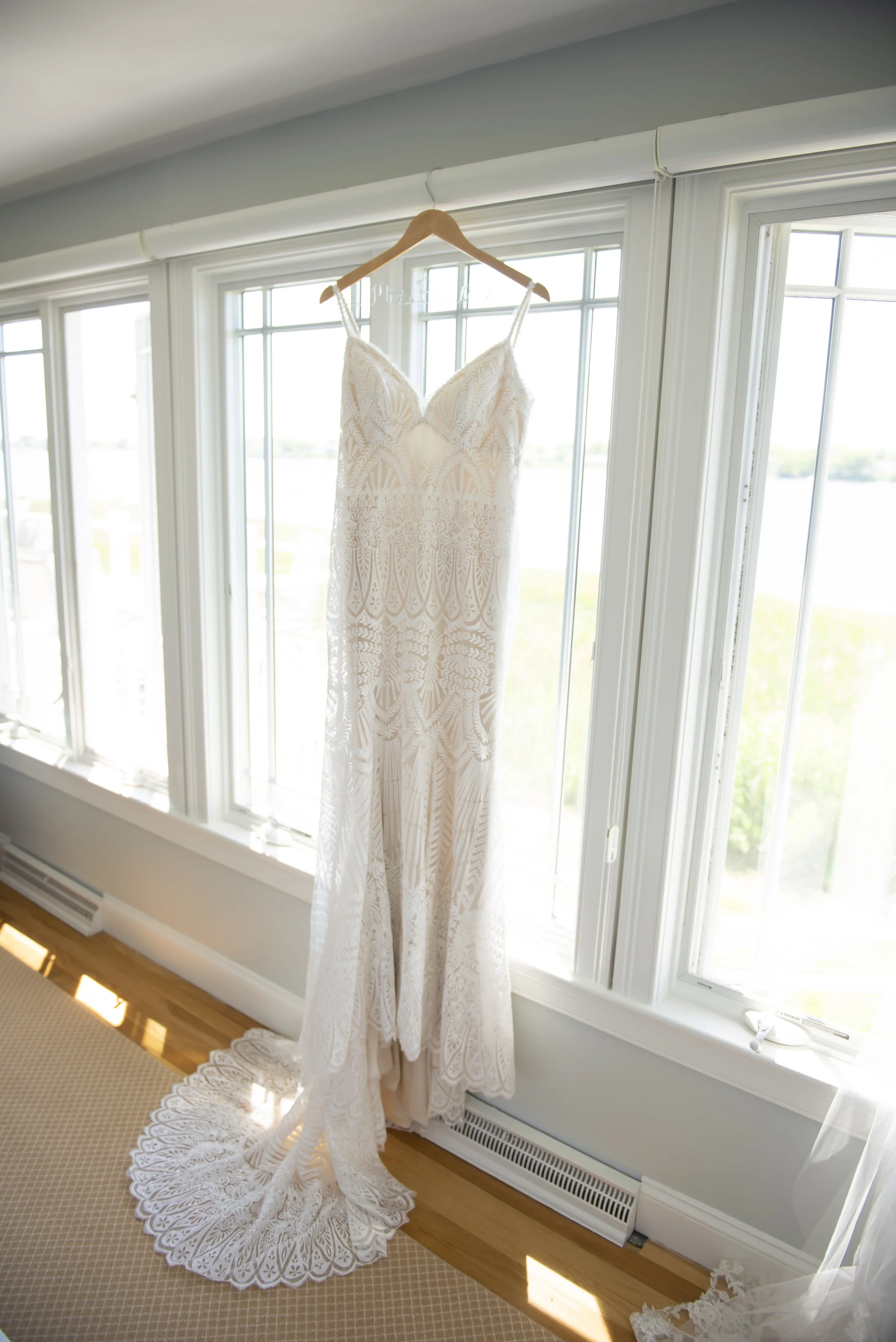 A white lace wedding dress hanging on a wooden hanger in front of large windows with sunlight streaming in.