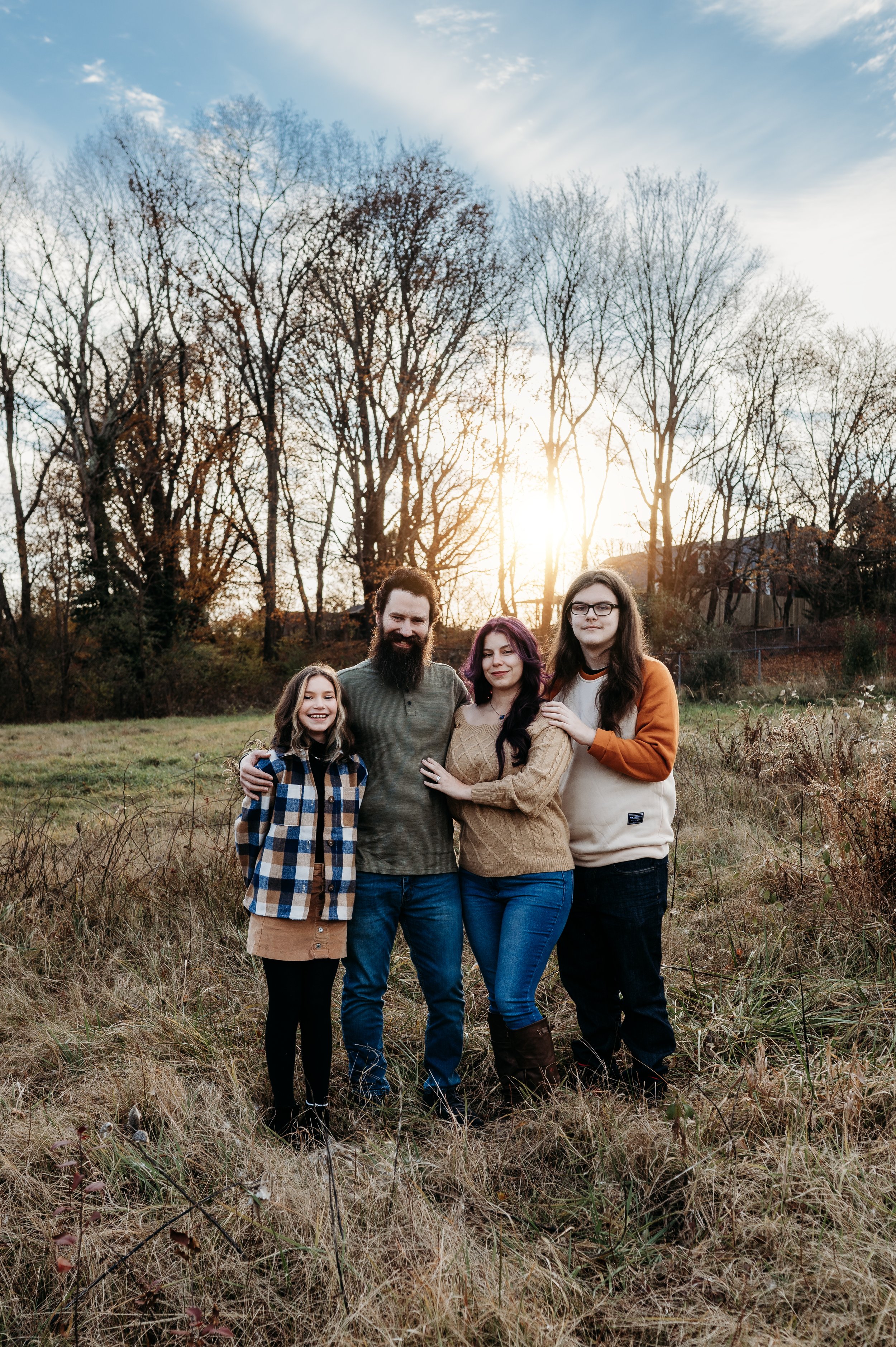 A family of four standing together outdoors in a grassy field with trees and the setting sun in the background, smiling at the camera.