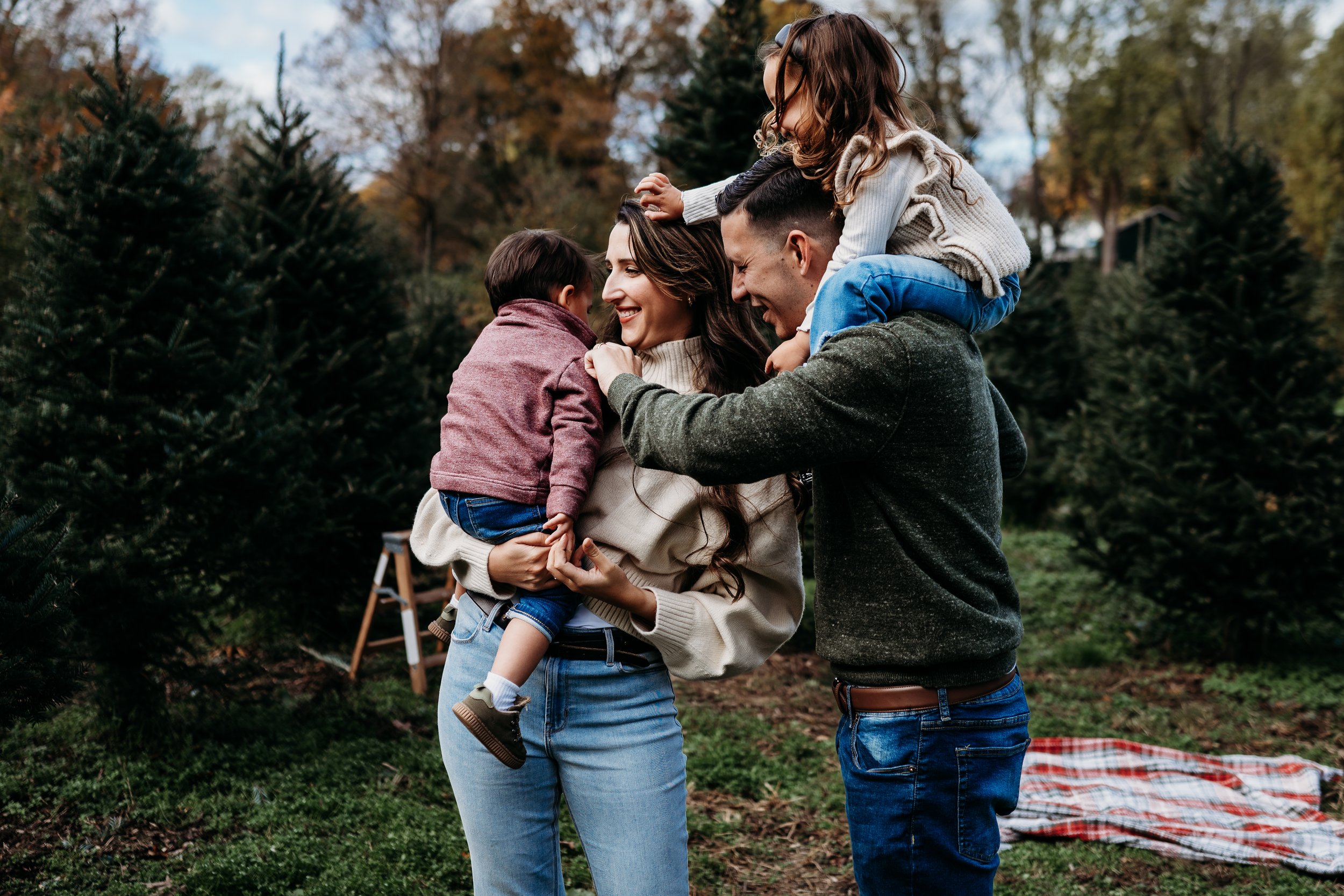A happy family of four outdoors during autumn, with the parents smiling and holding their two children, surrounded by evergreen trees.
