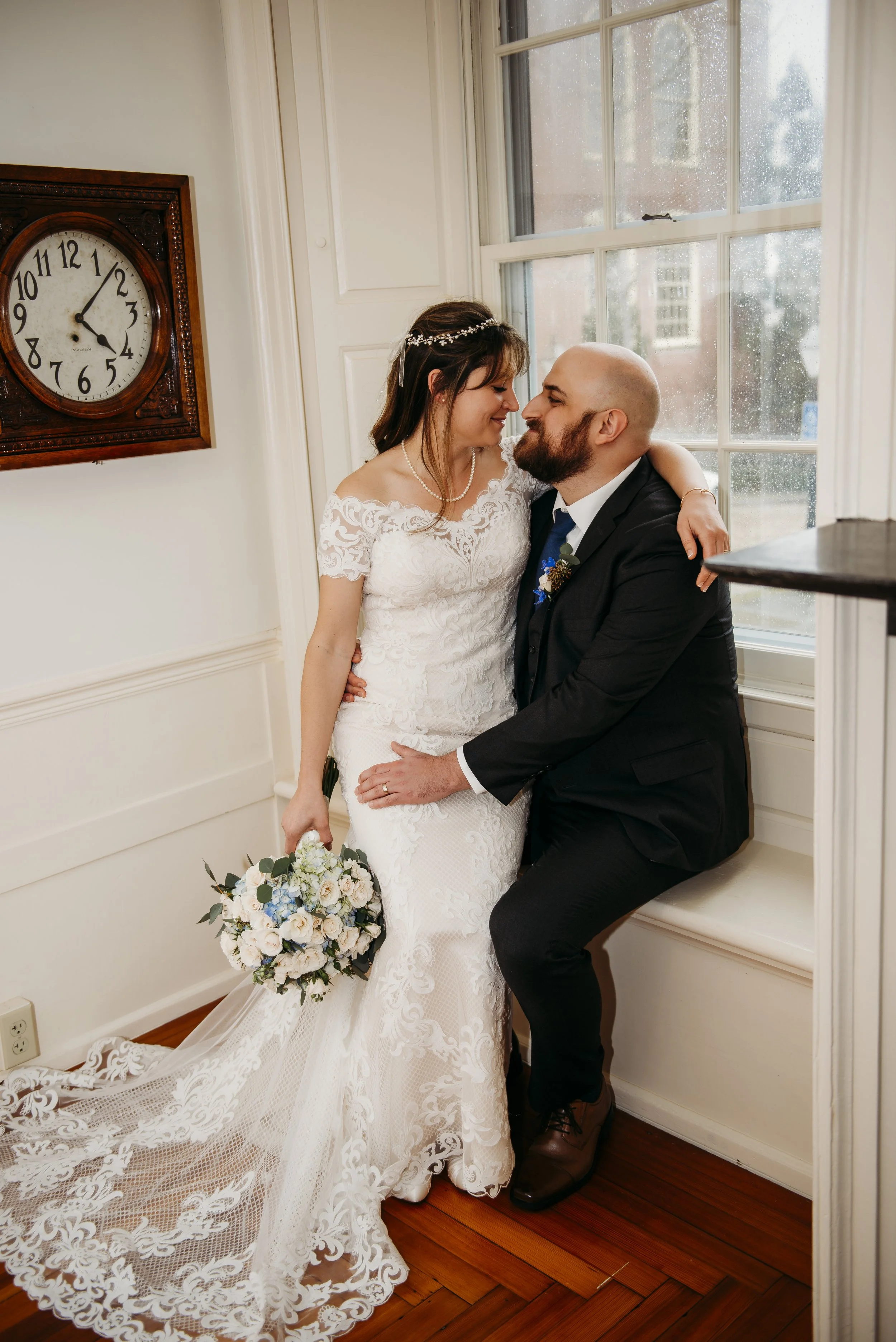 A bride and groom share an intimate moment indoors near a window, with a vintage clock on the wall behind them.