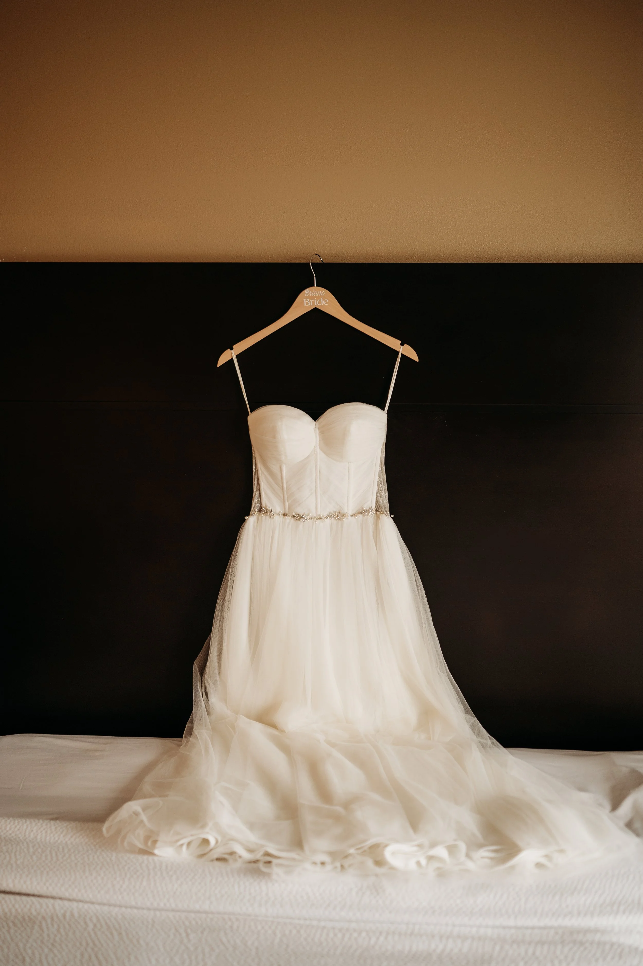 Elegant white wedding dress hanging on a bedside table with a black headboard in the background.