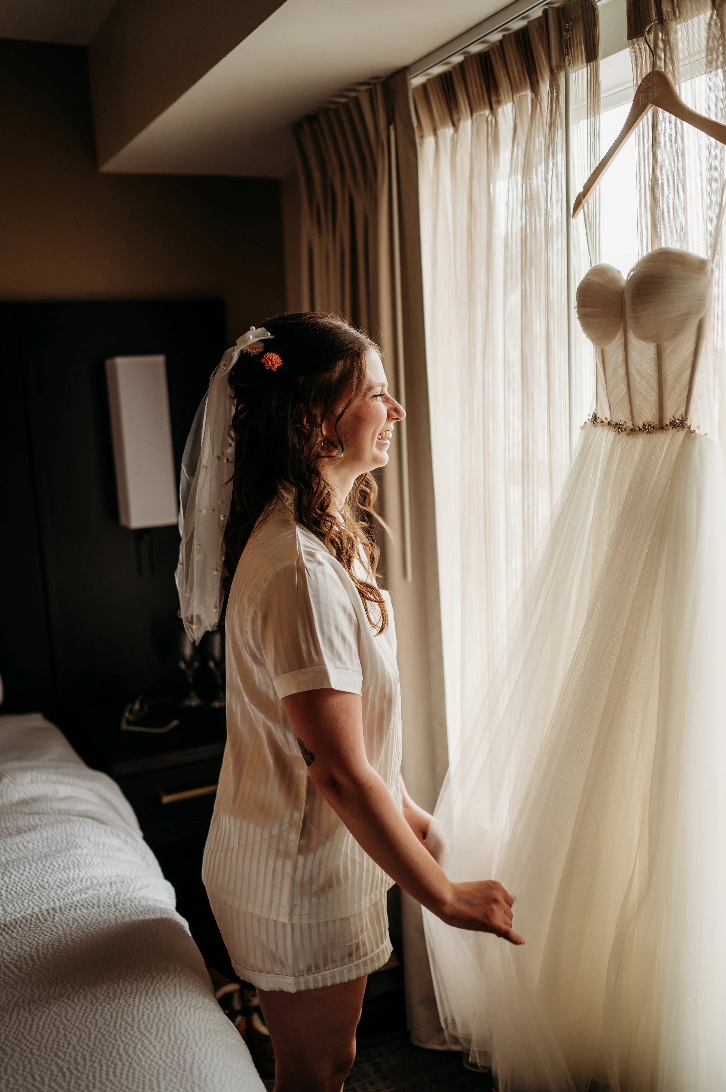 A woman with a veil and floral hair accessories looks happily at a wedding dress hanging by a window. She is smiling and holding the curtain.