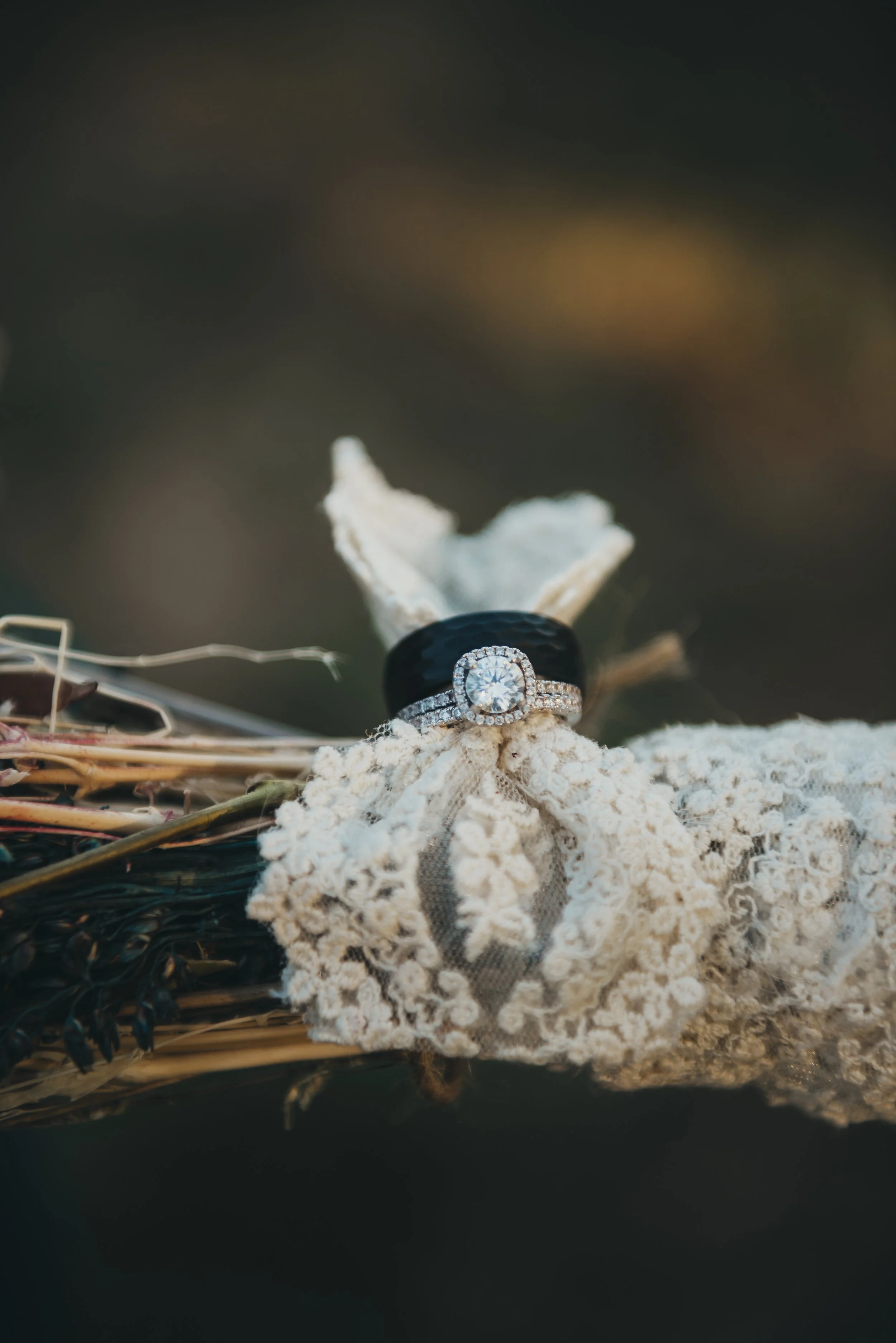 A diamond engagement ring and wedding band resting on a white fabric, with a black rubber ring behind them, in a rustic basket with dried flowers and twigs, outdoors at sunset.