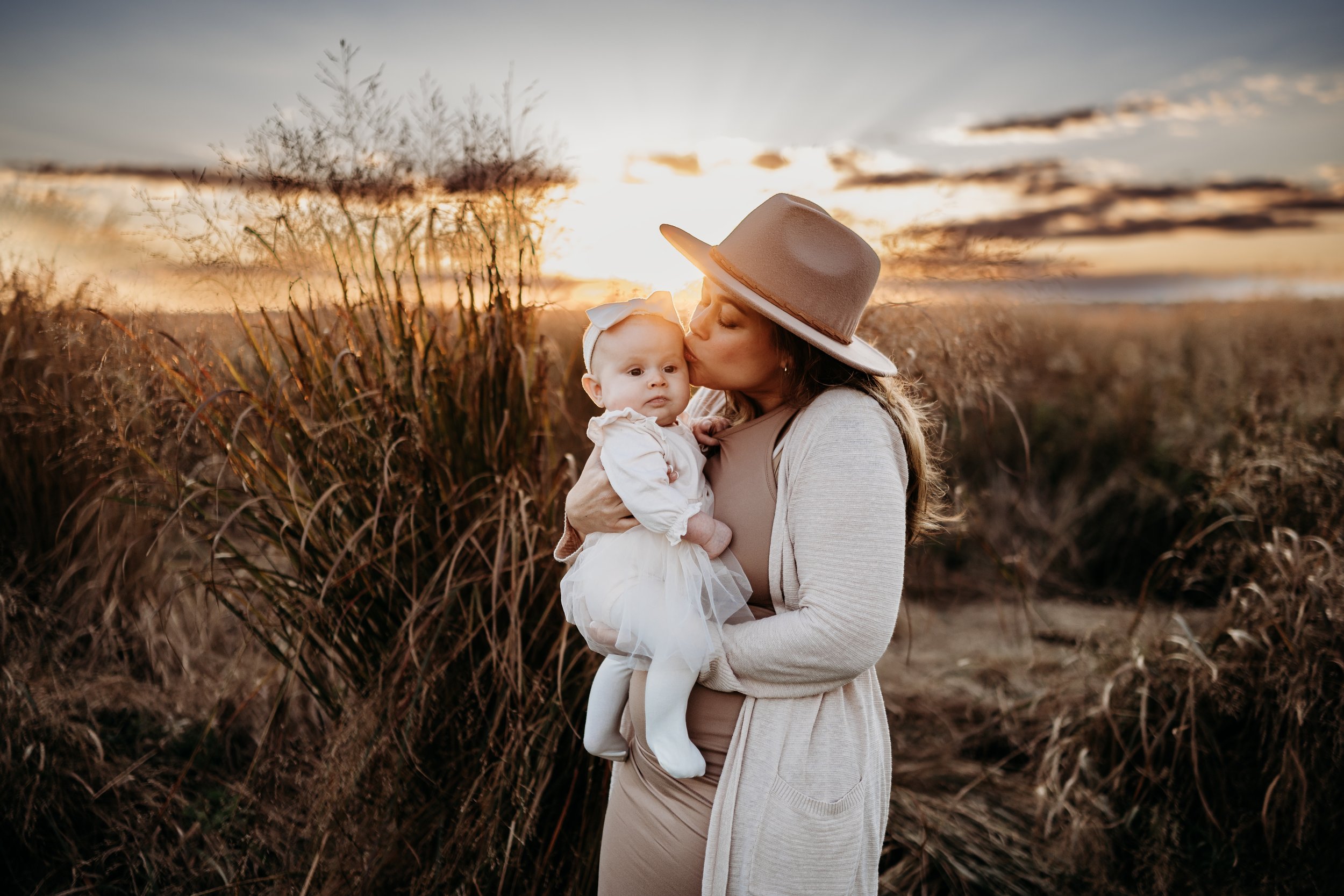 A woman holding a baby girl outdoors during sunset, with tall grasses in the background and a partly cloudy sky.