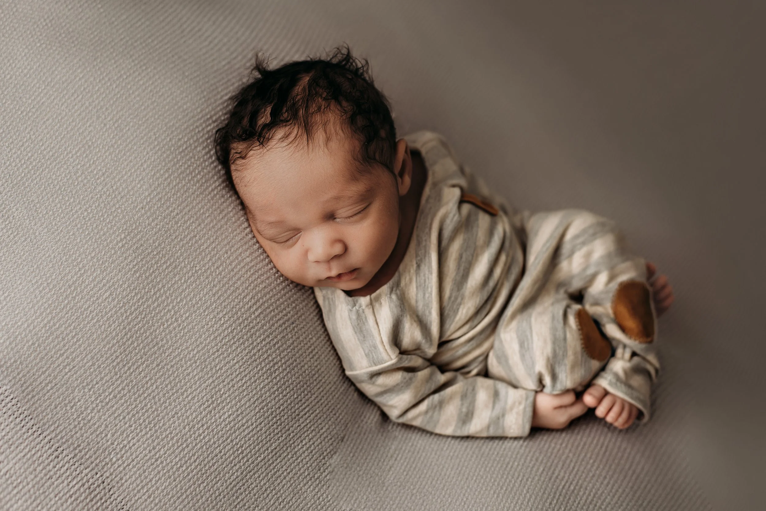 A sleeping newborn baby with dark curly hair, wearing a striped beige and brown outfit, lying on a textured gray surface.
