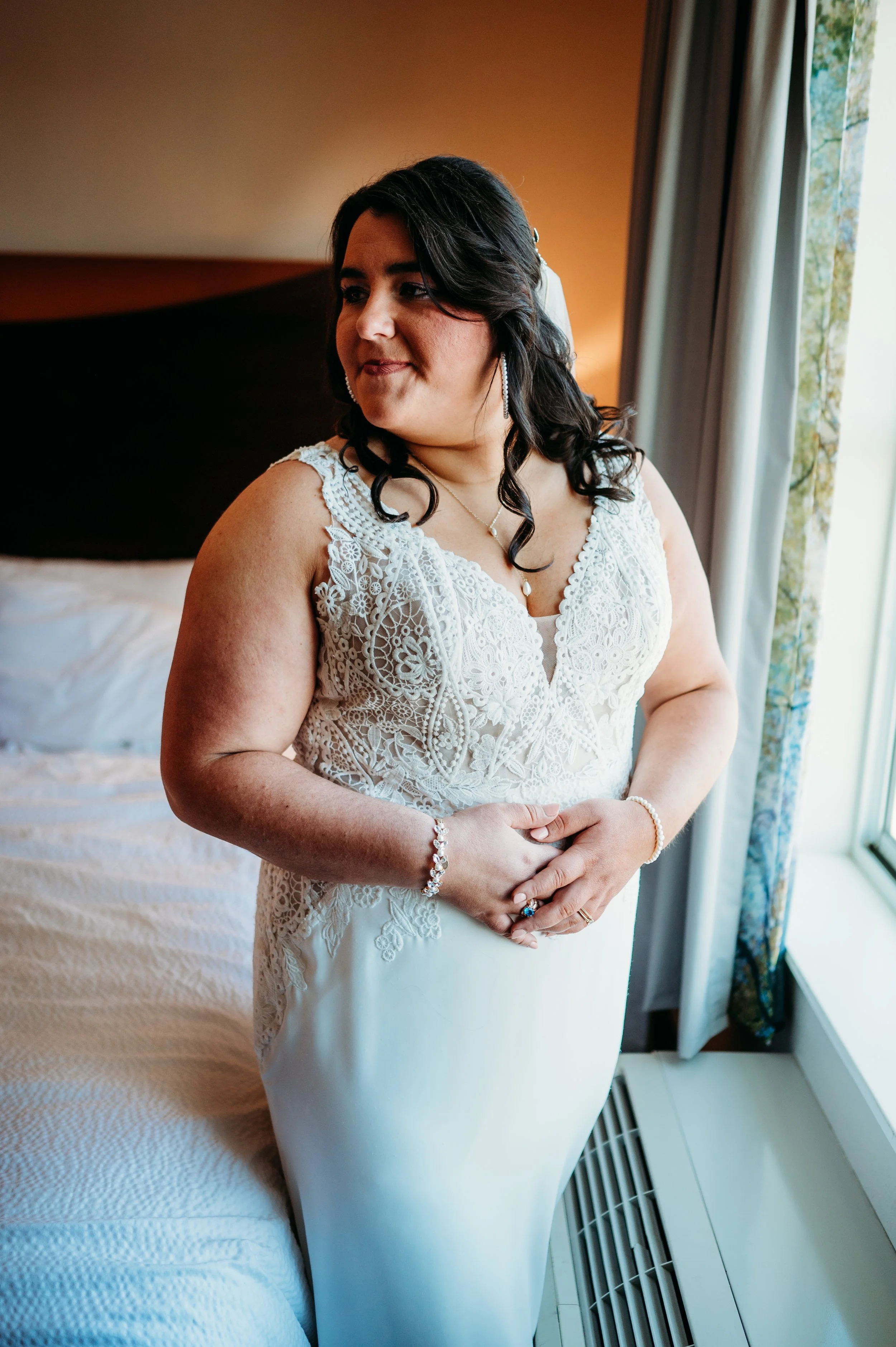 A woman in a white lace dress standing by a window, looking to her left, framed by curtains, with a bed and headboard in the background.