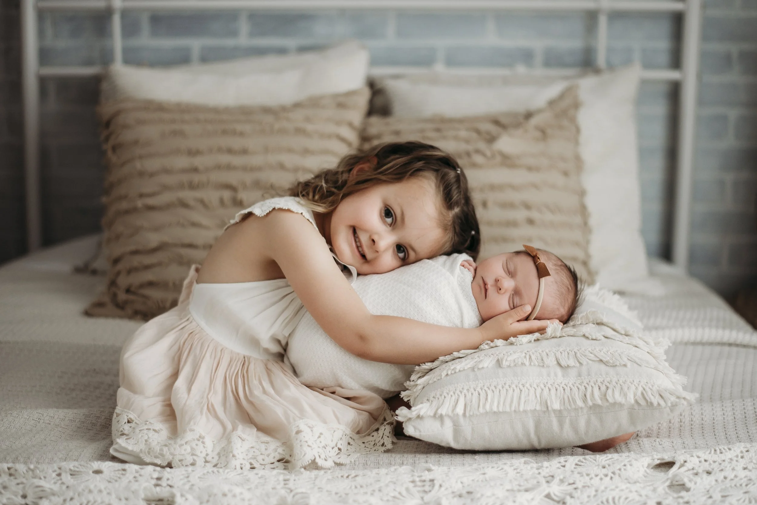 A young girl with long hair and a dress cuddles her newborn sister, who is sleeping on her side on a bed with pillows and a blanket.
