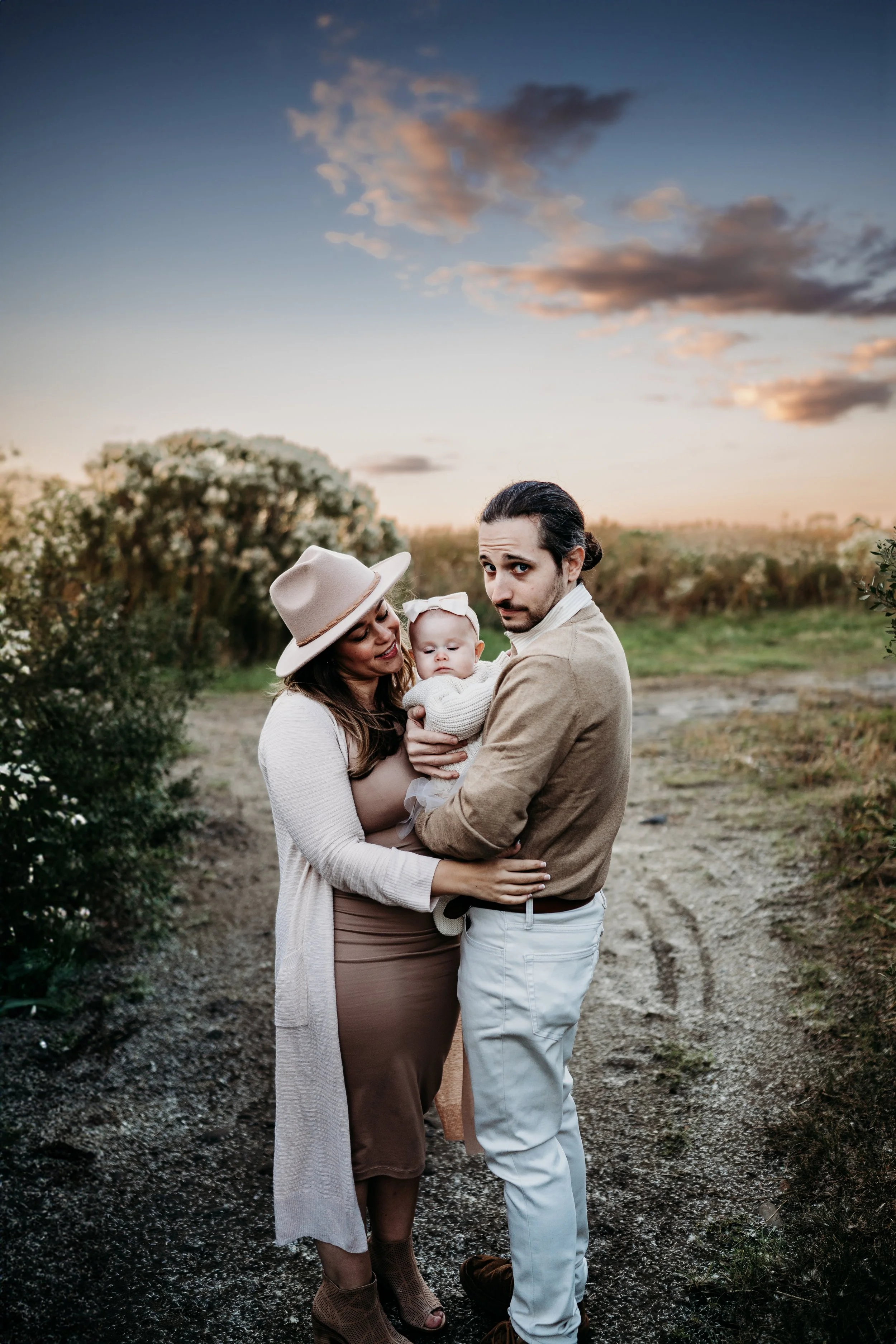 A family of three, a woman, a man, and a baby, standing outdoors on a dirt path at sunset, with greenery and clouds in the sky behind them.