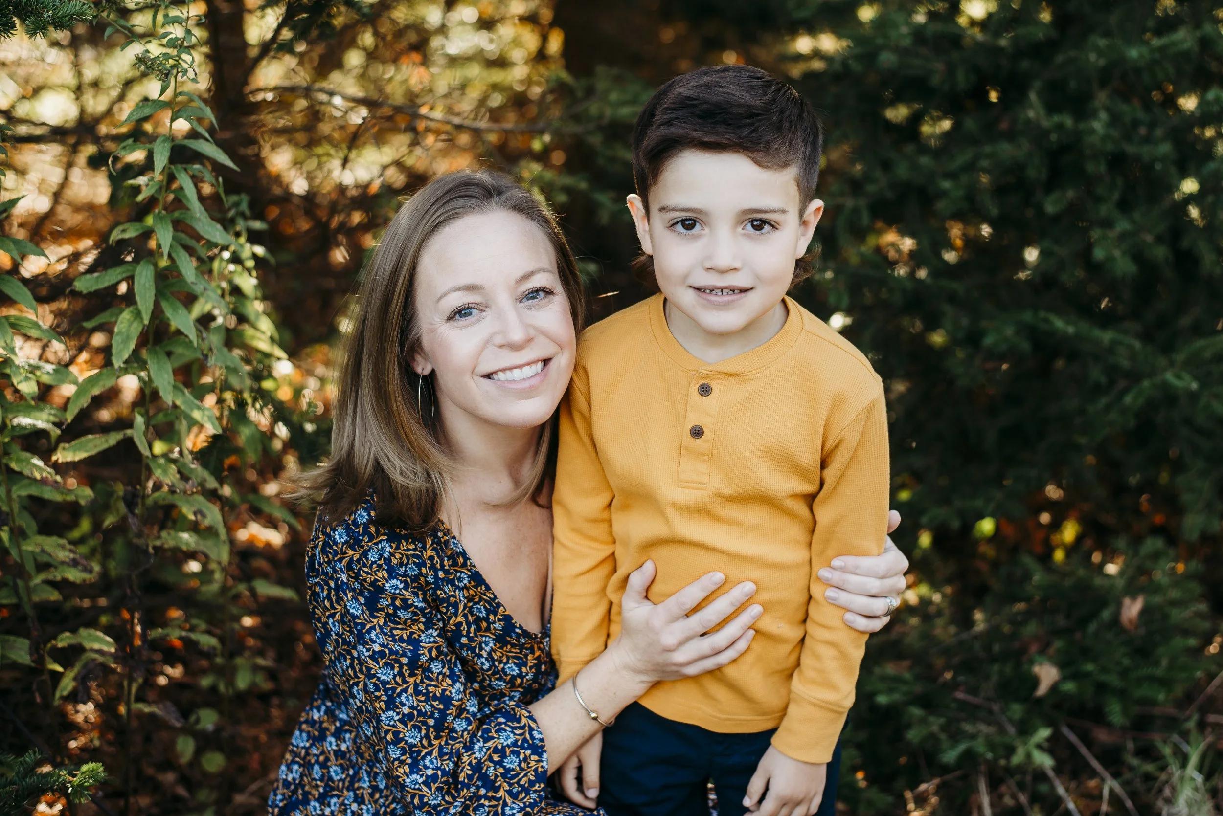 A smiling woman with shoulder-length brown hair in a floral dress is hugging a young boy with dark brown hair wearing a mustard yellow shirt. They are outdoors with green foliage and brown leaves in the background.