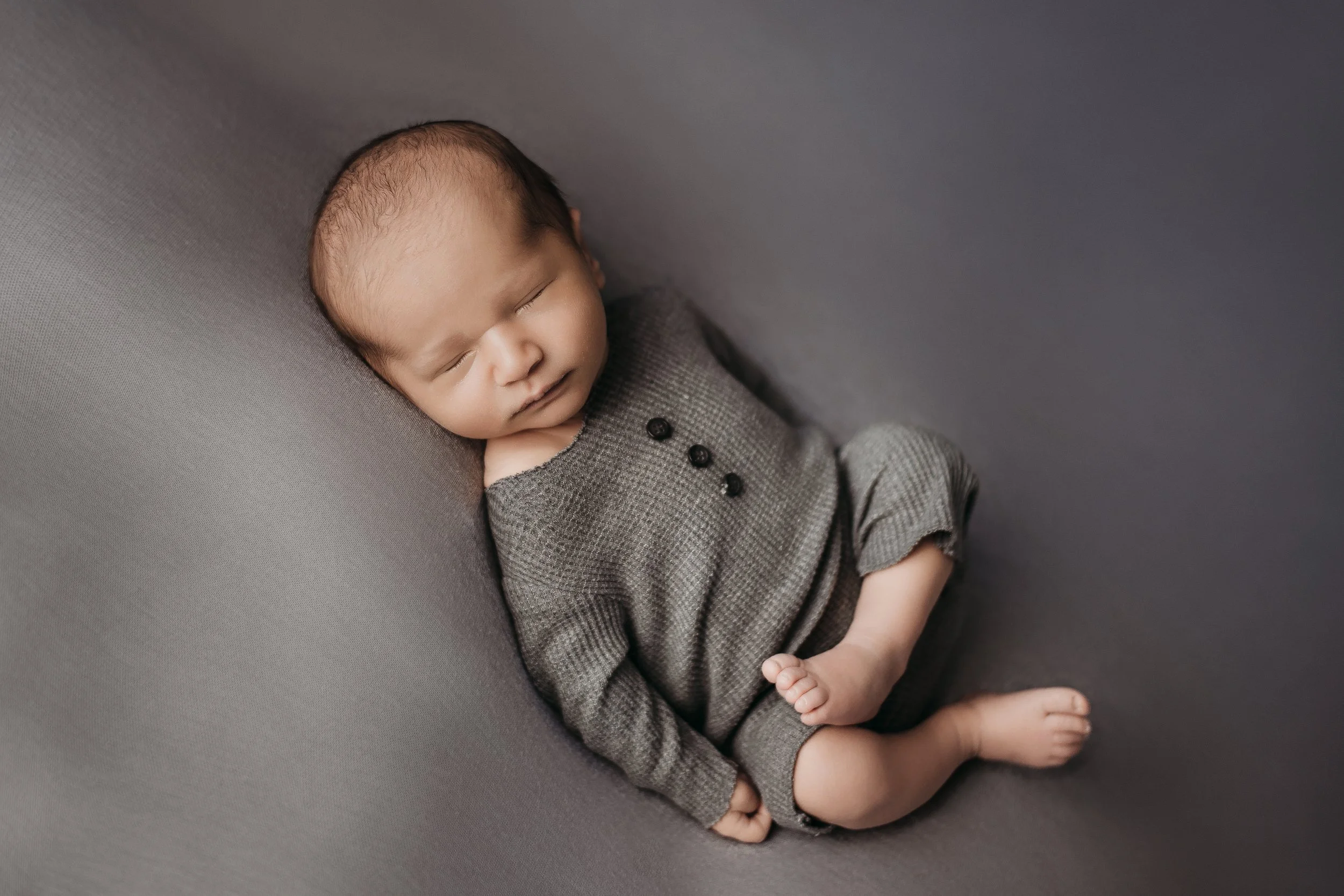 A sleeping baby with light skin, wearing a gray long-sleeve outfit, resting on a soft gray surface.