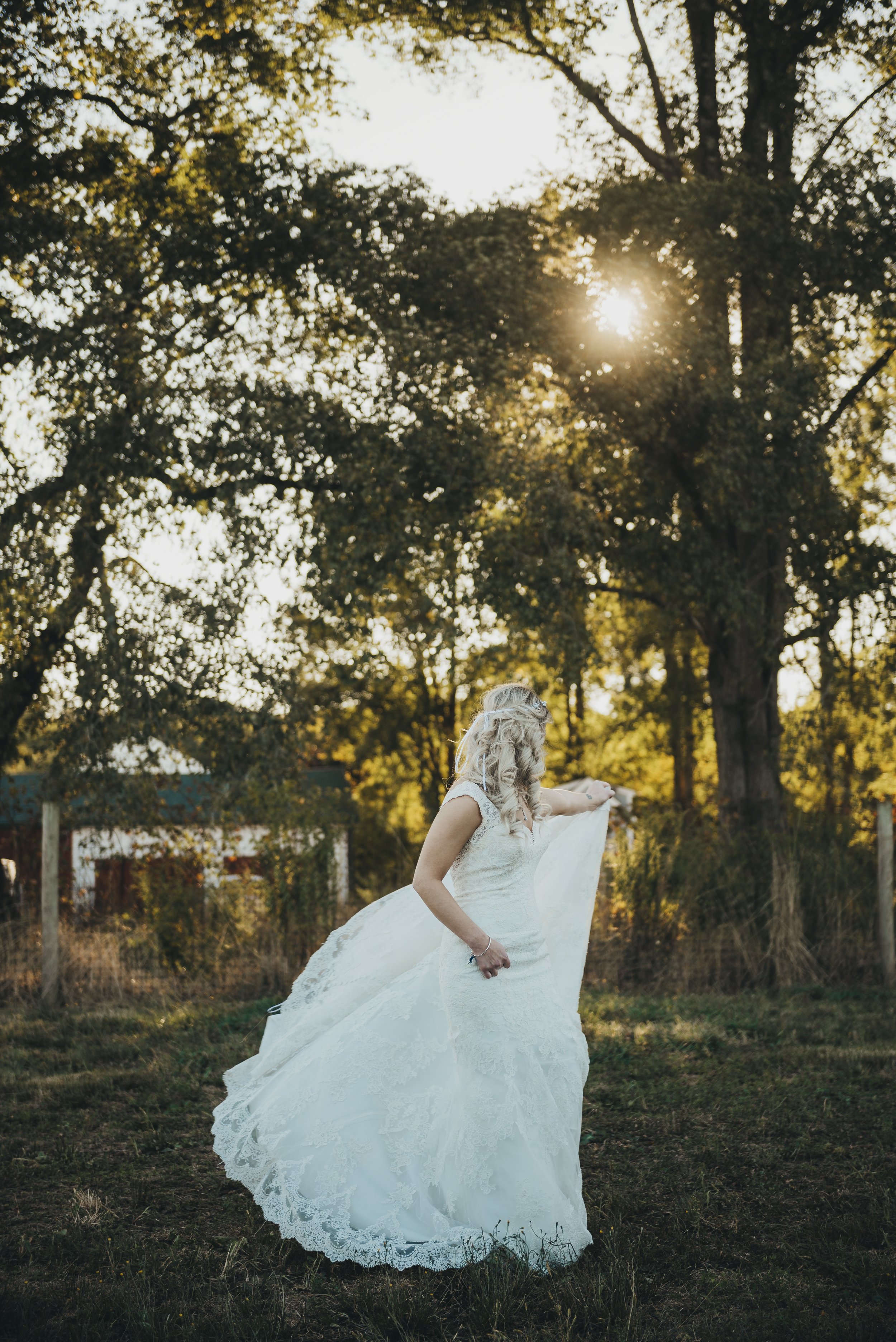 Bride in a white wedding gown outdoors during sunset, holding up part of her dress, with trees in the background.