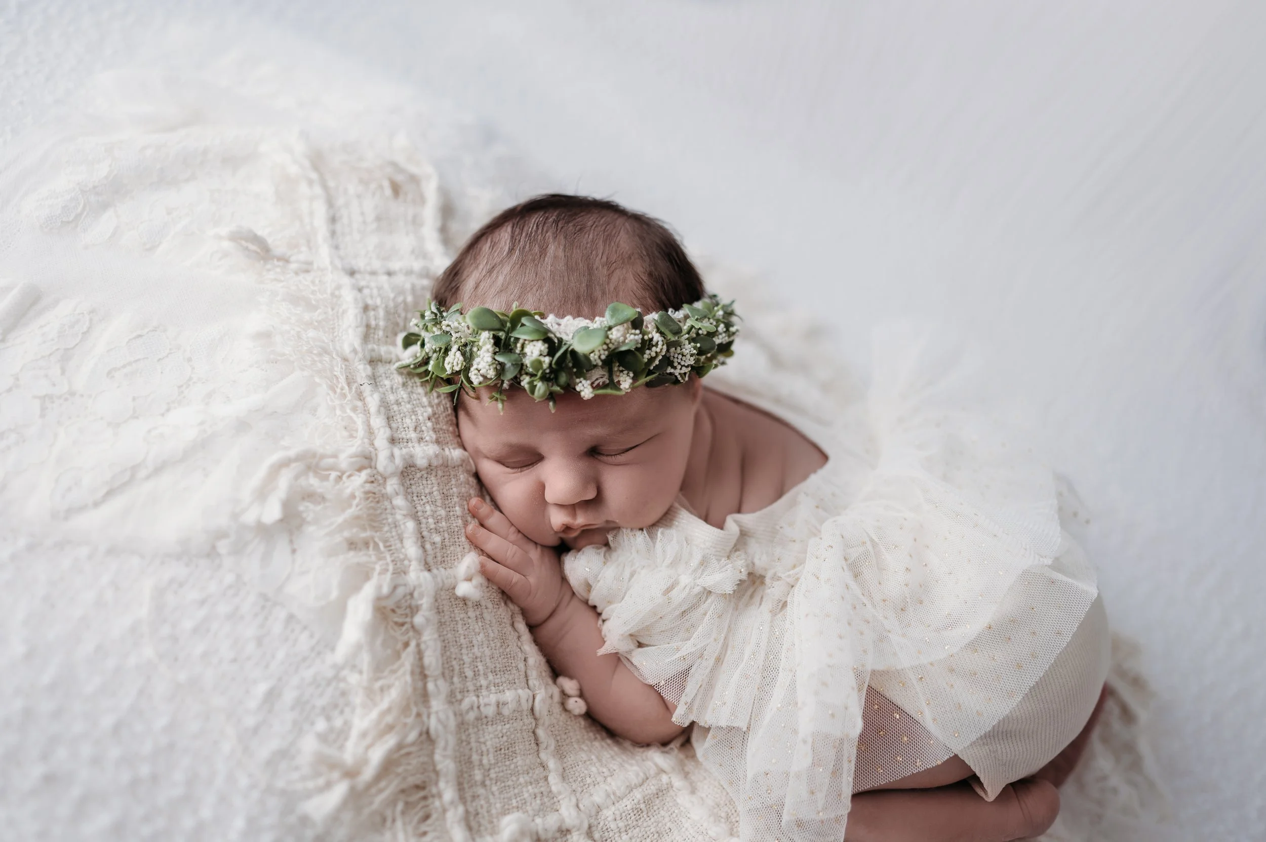 A sleeping newborn baby with a floral headband, dressed in a lacy outfit, lying on a soft blanket.