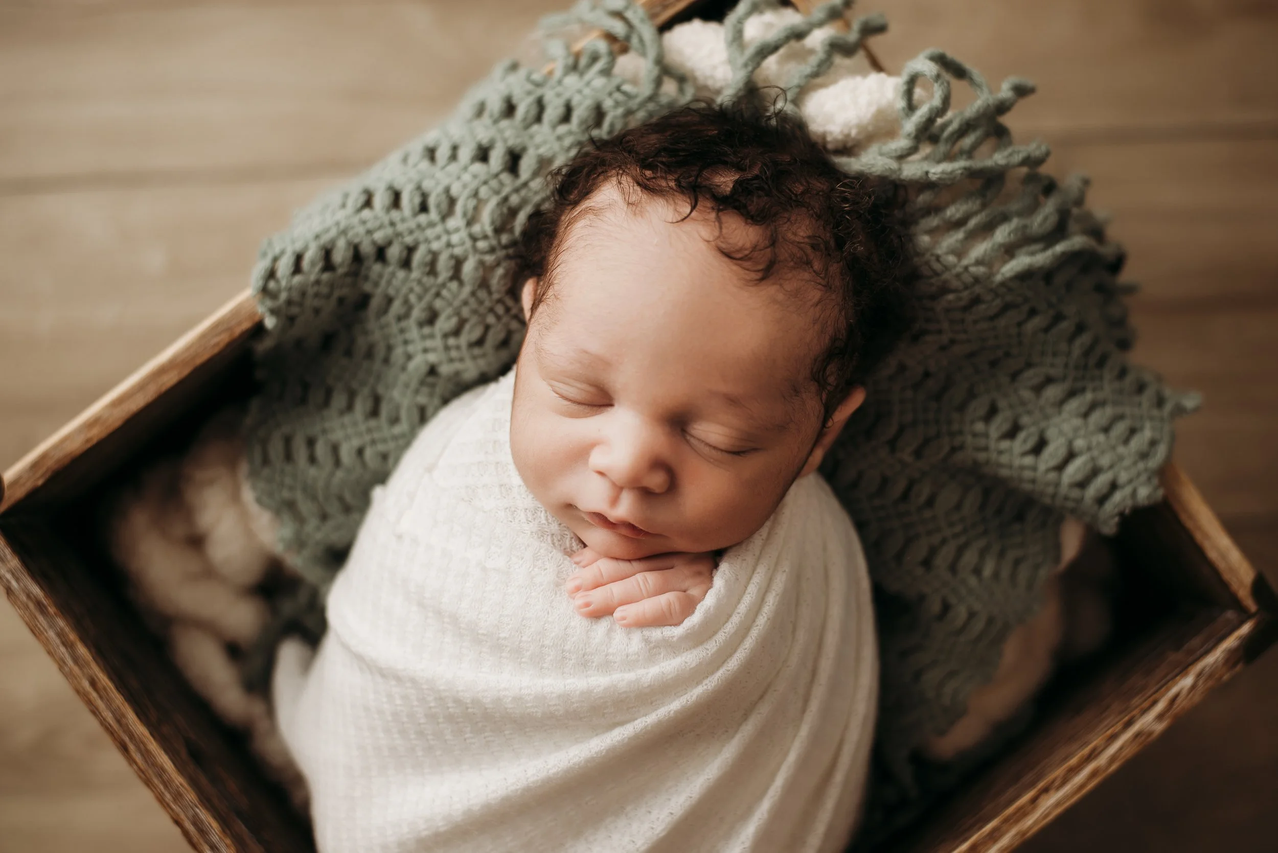 A sleeping newborn baby wrapped in a white blanket, resting on a knit blanket inside a wooden basket.