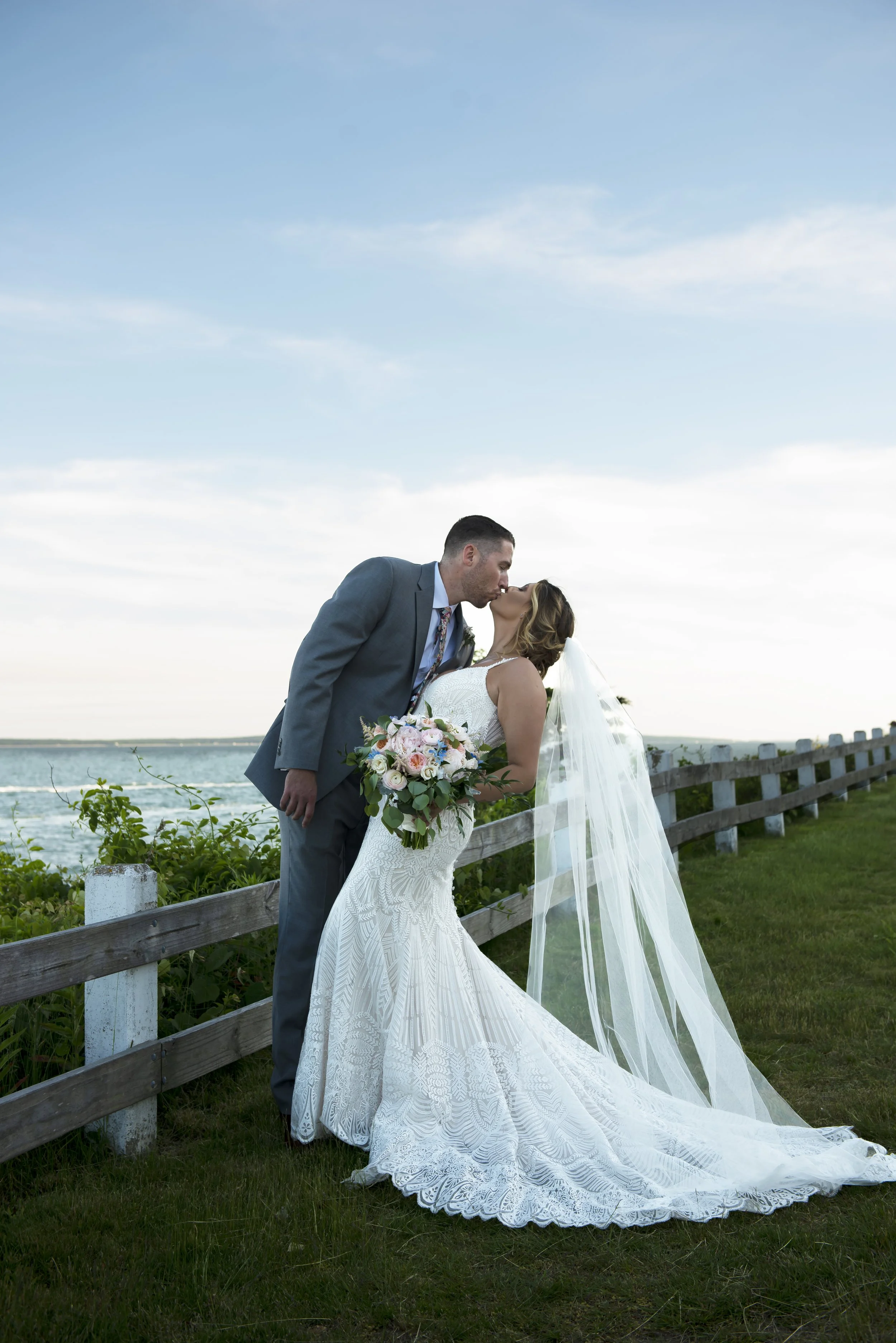 A newlywed couple sharing a kiss by a wooden fence near water, with the bride in a white lace wedding gown holding a bouquet, and the groom in a blue suit, during sunset or late afternoon.