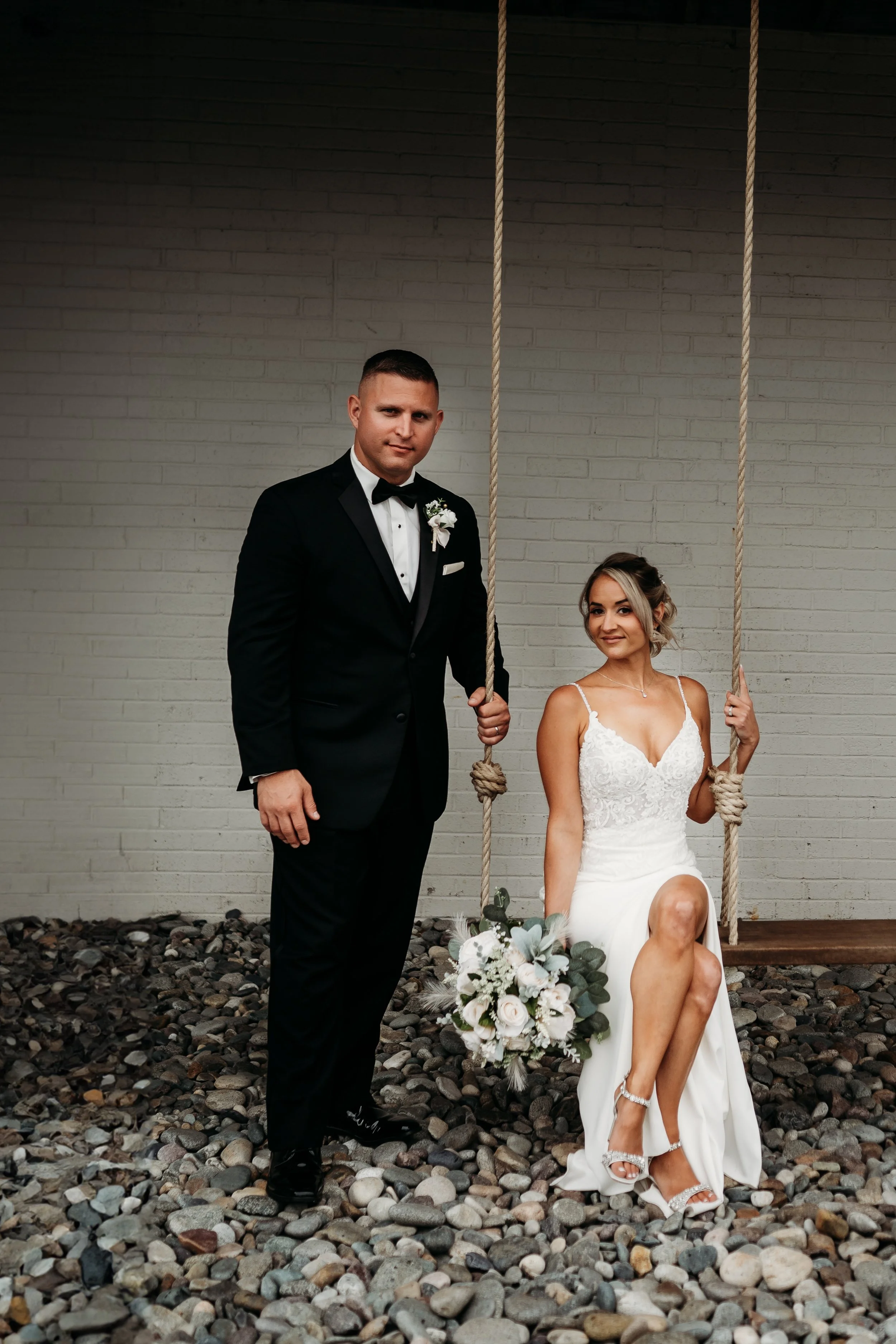 A bride in a white wedding dress sitting on a swing with a bouquet, and a groom in a black tuxedo standing next to her, against a white brick wall.