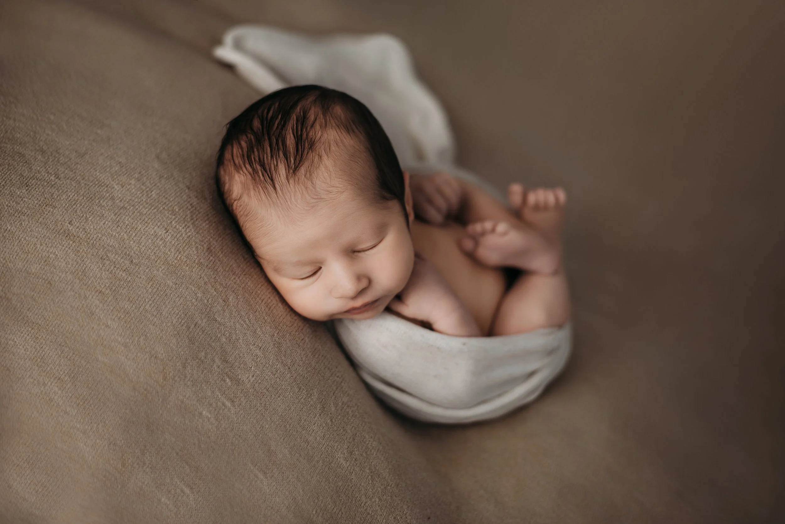 Newborn baby peacefully sleeping on a brown couch, wrapped in a light-colored blanket.