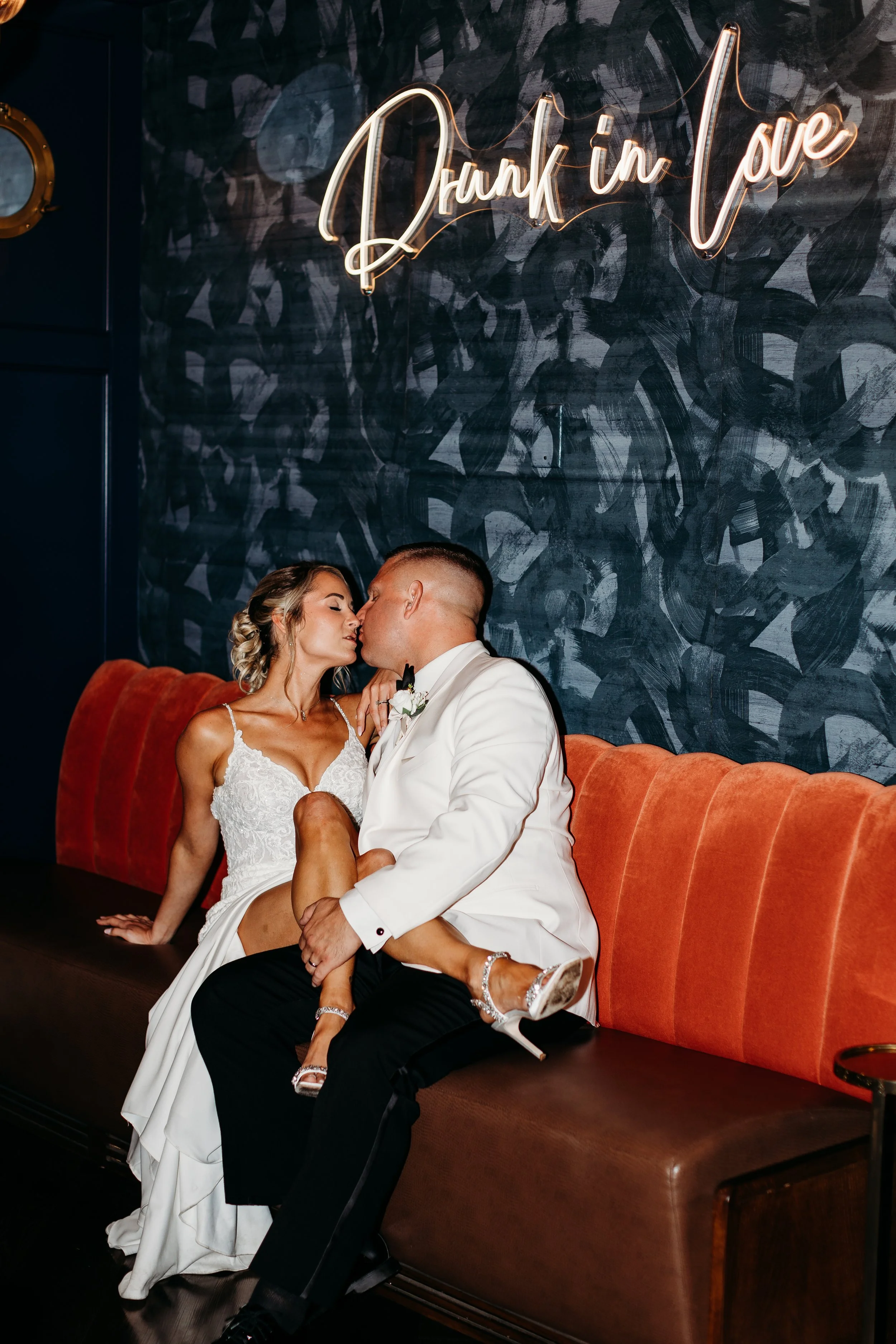 A bride and groom kiss on a sofa in a modern venue with a dark wall background and neon sign that says 'Drunk in Love'.