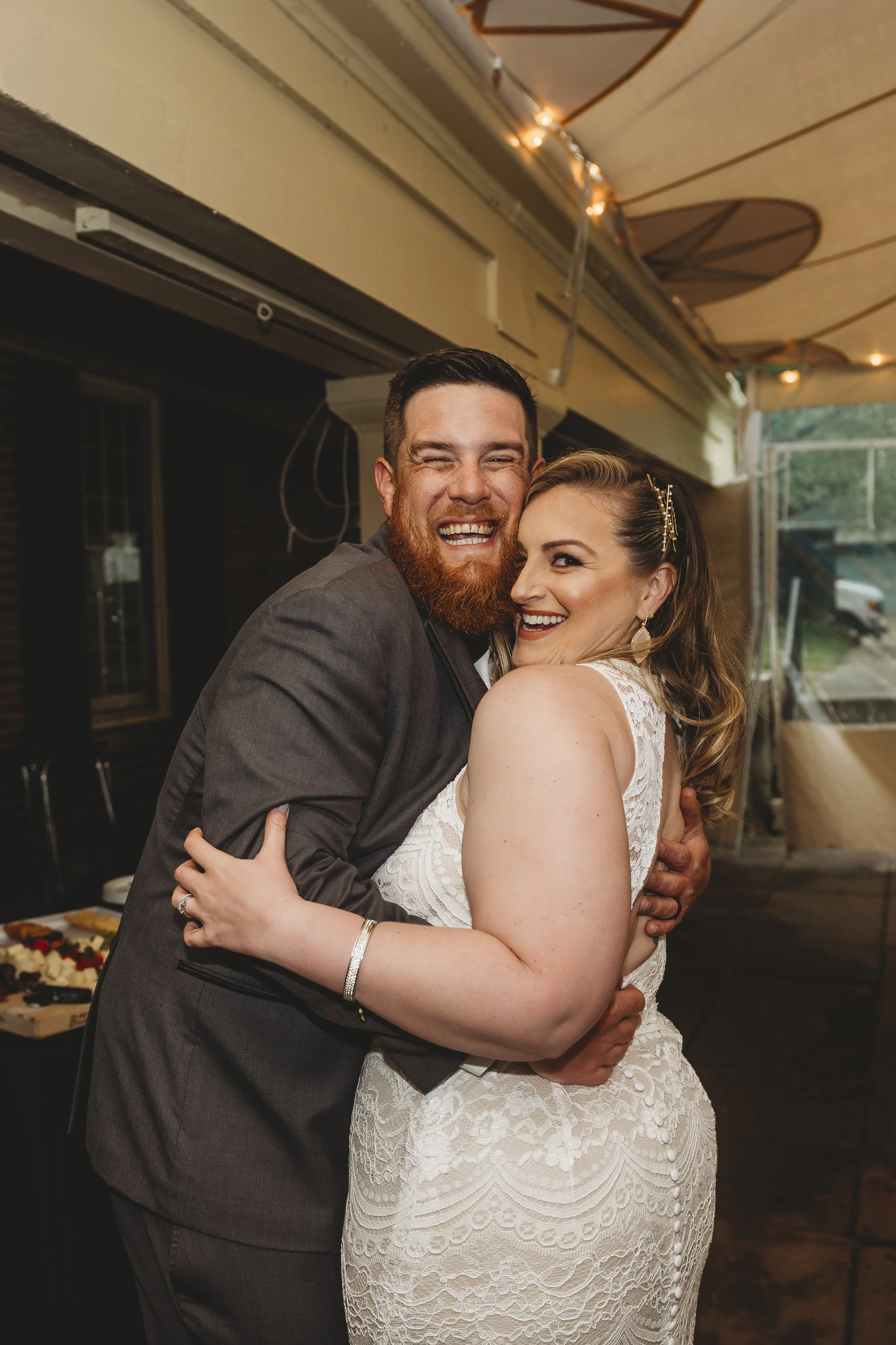A happy couple, dressed in wedding attire, hugging and smiling at a wedding reception. The bride is in a white lace dress, and the groom in a grey suit.
