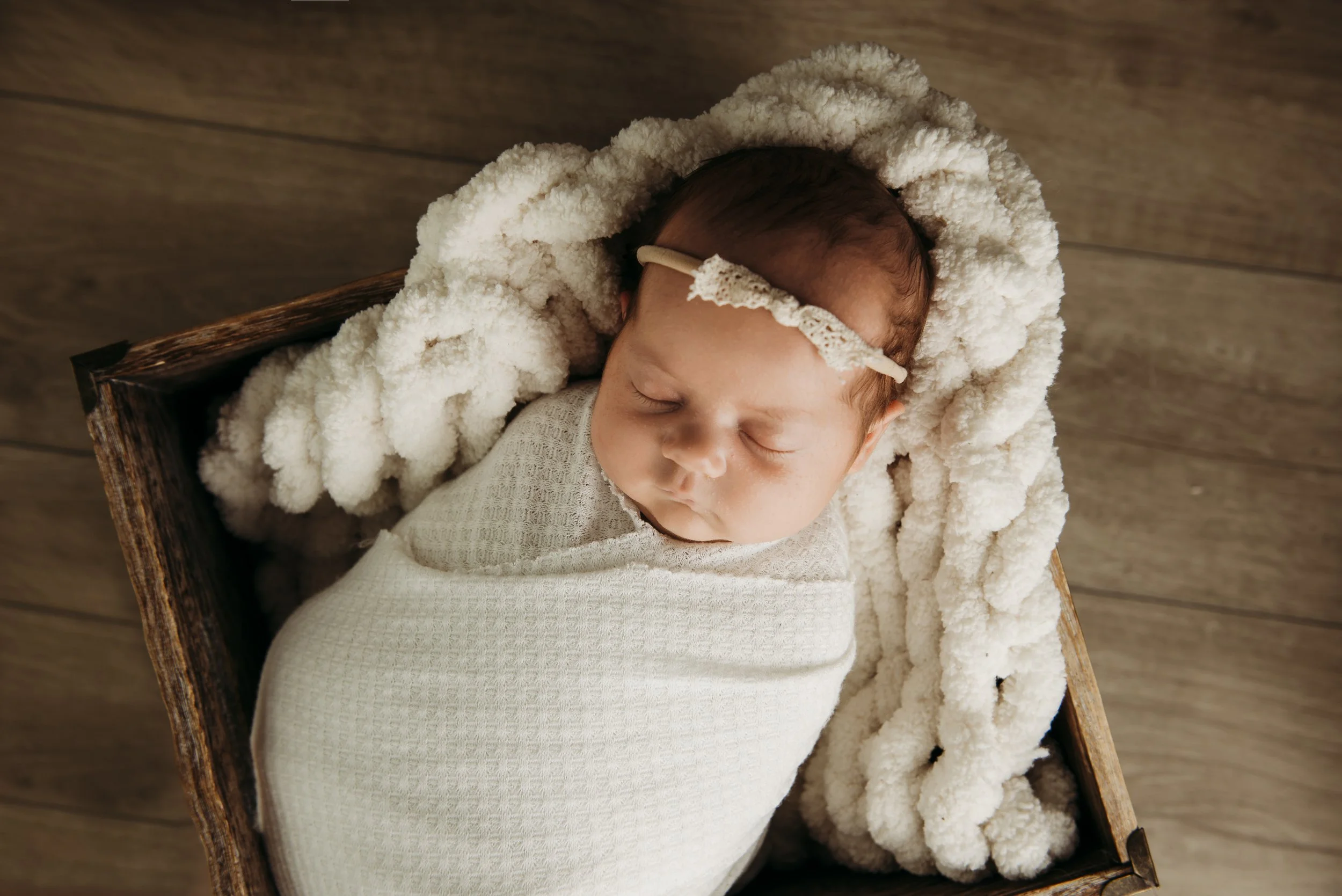 A sleeping newborn baby swaddled in a beige blanket, resting on a fluffy cream-colored blanket inside a wooden box, with a lace headband on her head, on a wooden floor.