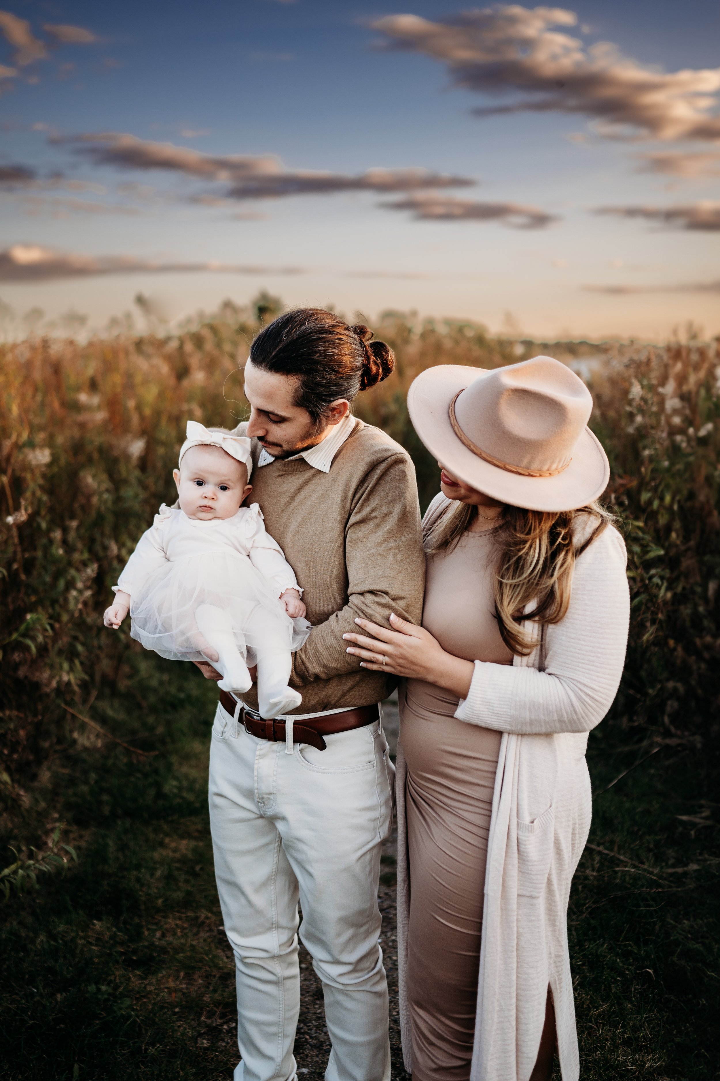 A family of three, including a man, woman, and a baby girl, standing outdoors at sunset among plants. The man is holding the baby who is dressed in white, and the woman is touching the man's arm. The woman is wearing a large beige hat and a beige dre