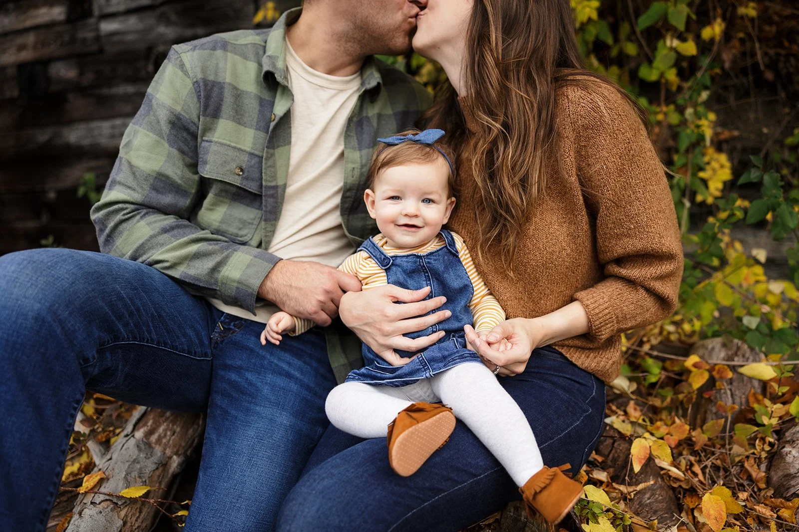 A family of three sitting on a log outdoors with Fall leaves at their Seneca Lake family session.