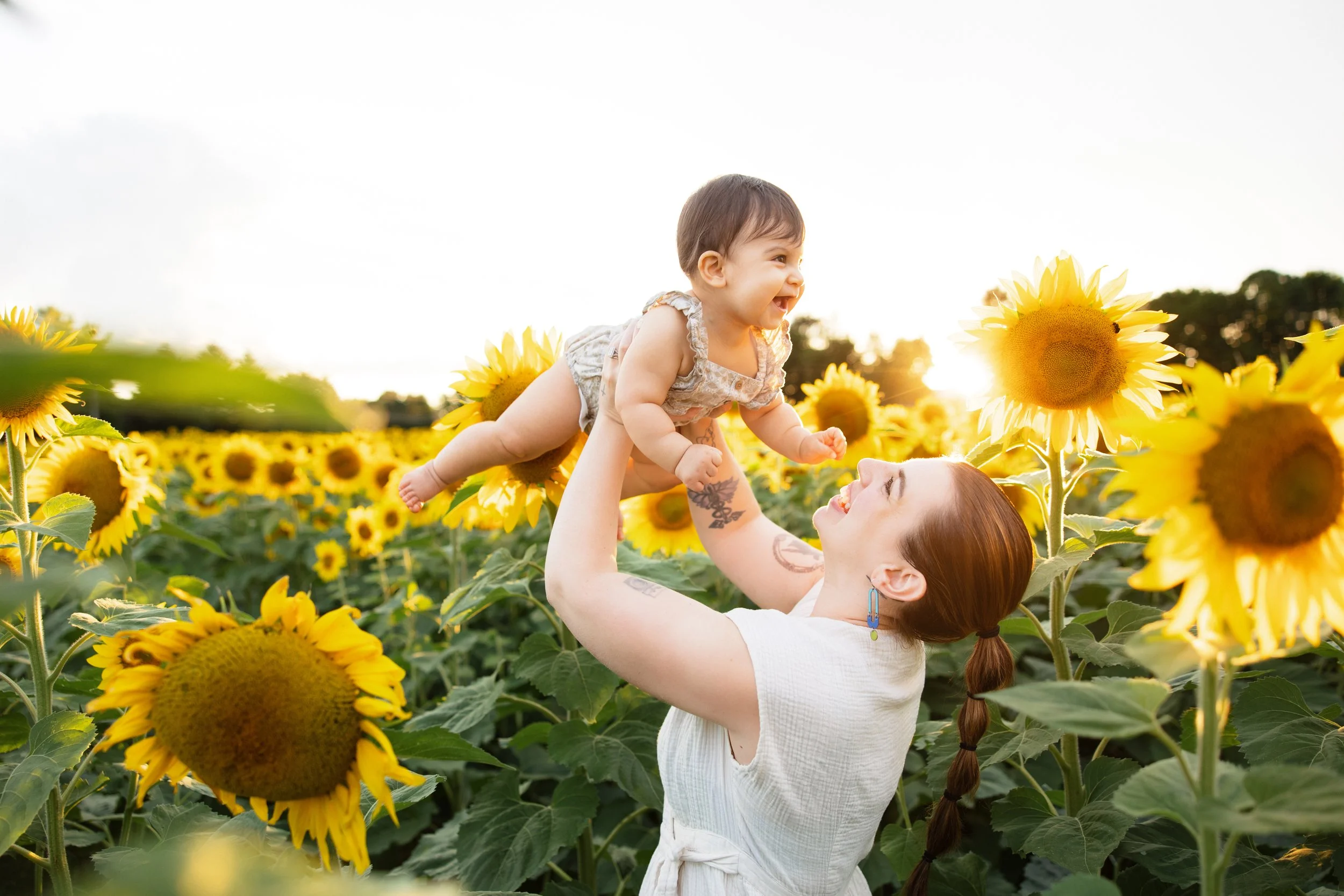 A mother with long brown hair in a braid and tattoos on her arm lifting a smiling baby girl in a dress in a sunflower field during sunset in Rochester, NY.